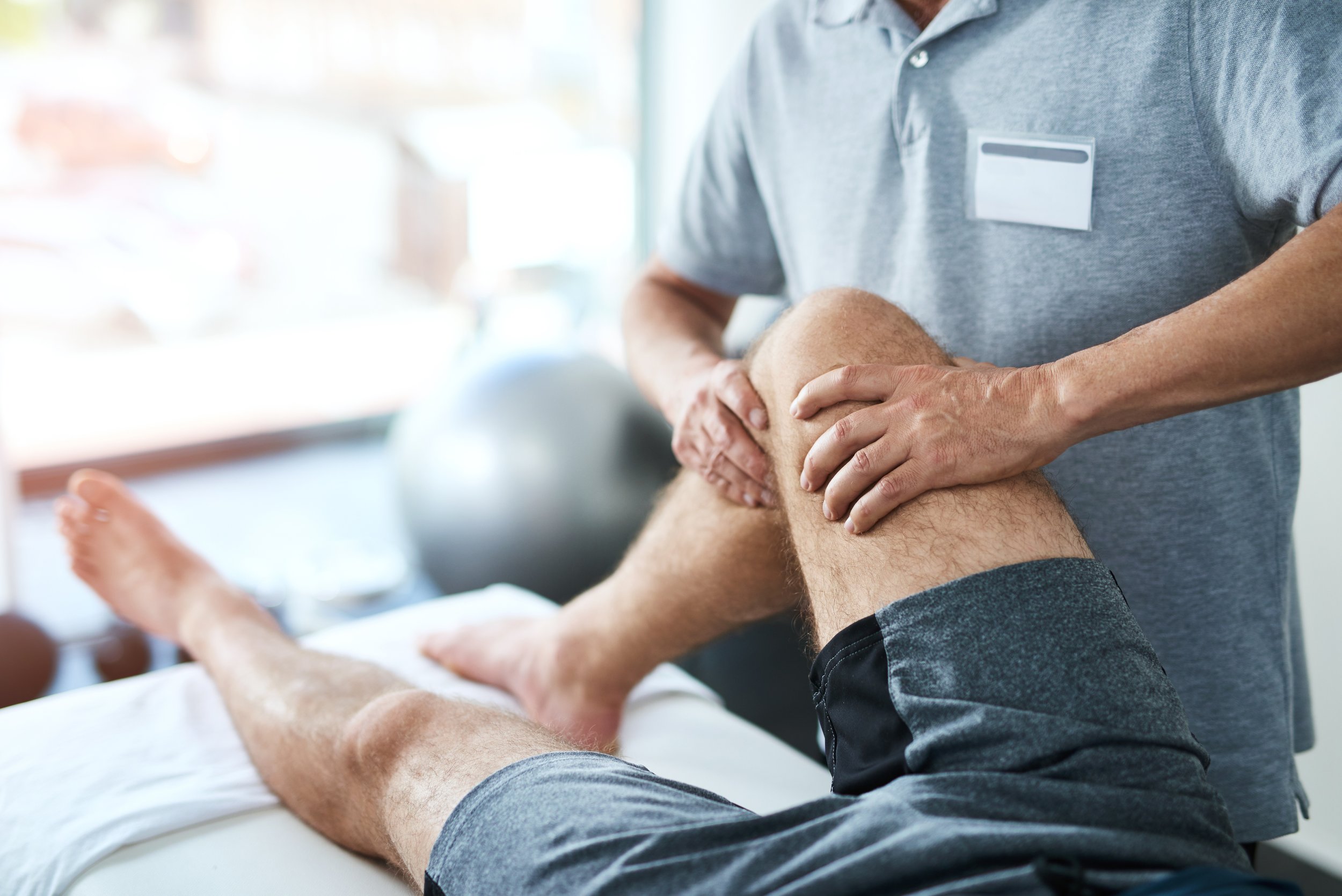 Einn Physiotherapeut macht Krankengymnastik mit einem Patienten, um das KNie wieder in Ordnung zu bringen. Der Patient liegt auf der Liege, das Knie ist angewinkelt.