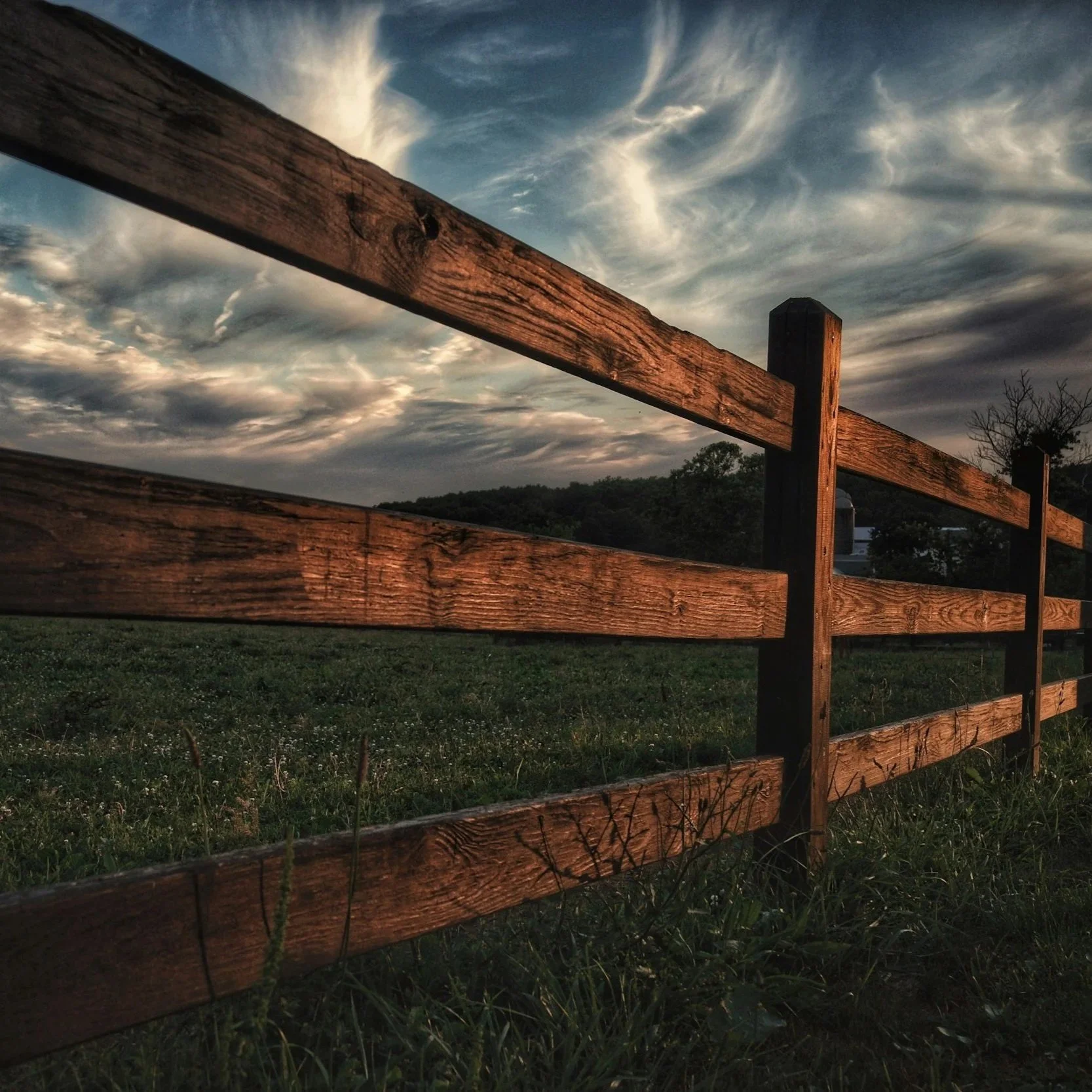 Ranch-style wooden fence in grassy field beneath cloudy sky, representing durability, weather impact, and benefits of correctly timed professional fence installation.