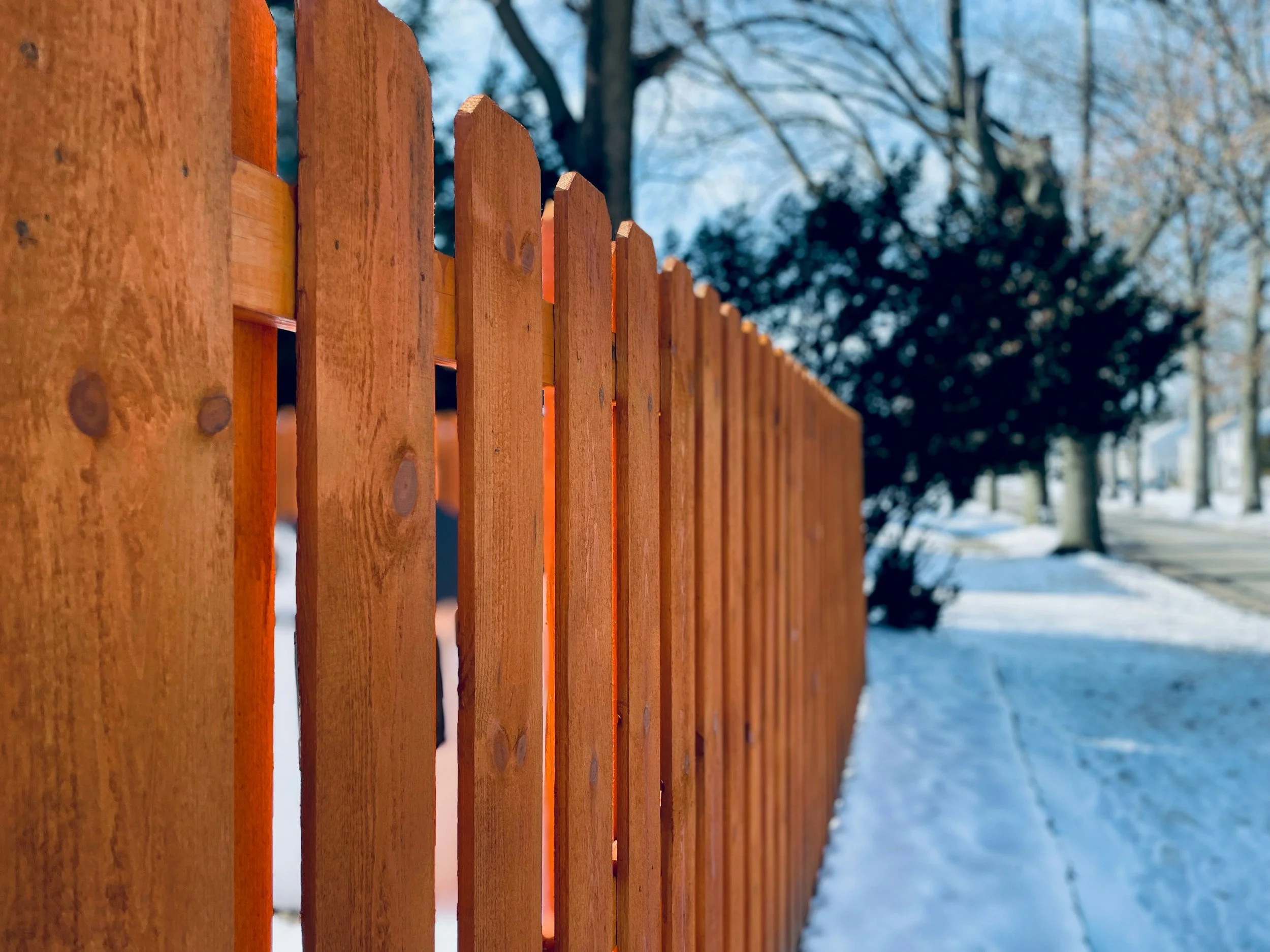 Close-up of a wooden picket fence along a snow-covered sidewalk in winter, with trees and houses in the background.