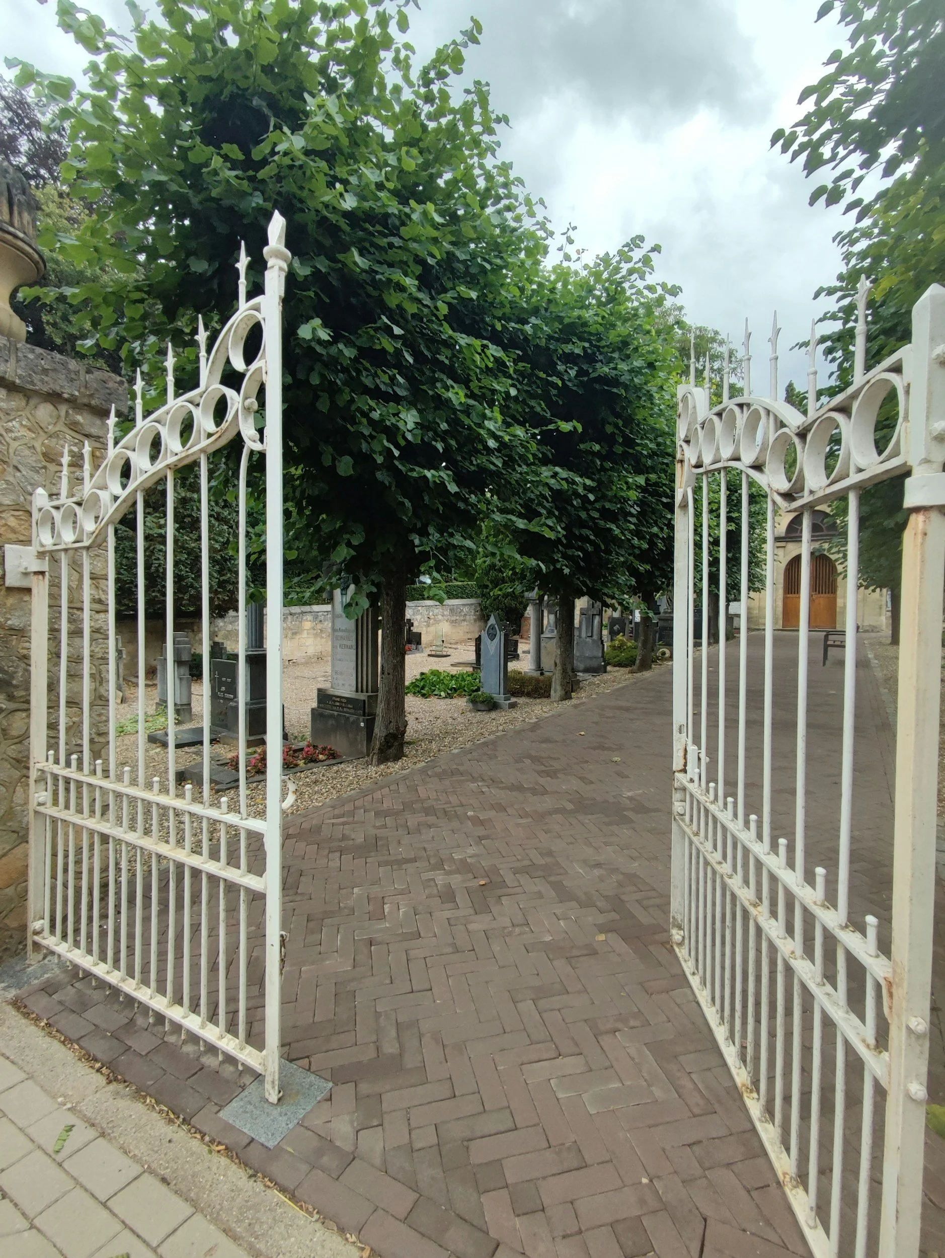 Open white metal gate leading into a small cemetery with headstones and trees
