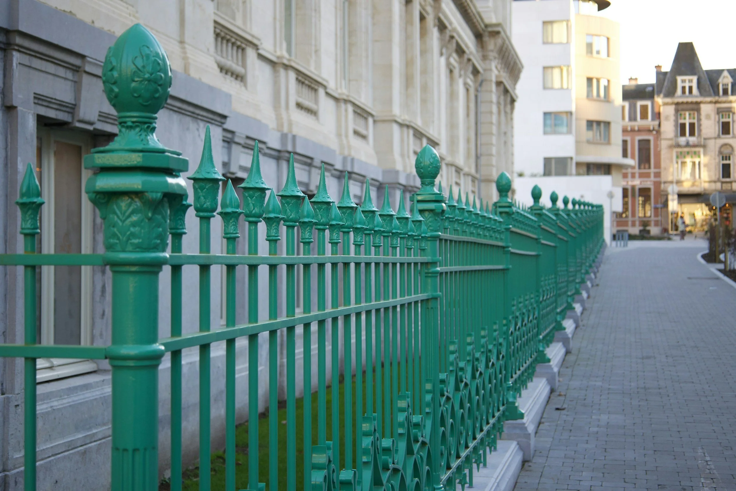 A street with a decorative teal metal fence in front of a white ornate building, with a sidewalk and other buildings in the background.