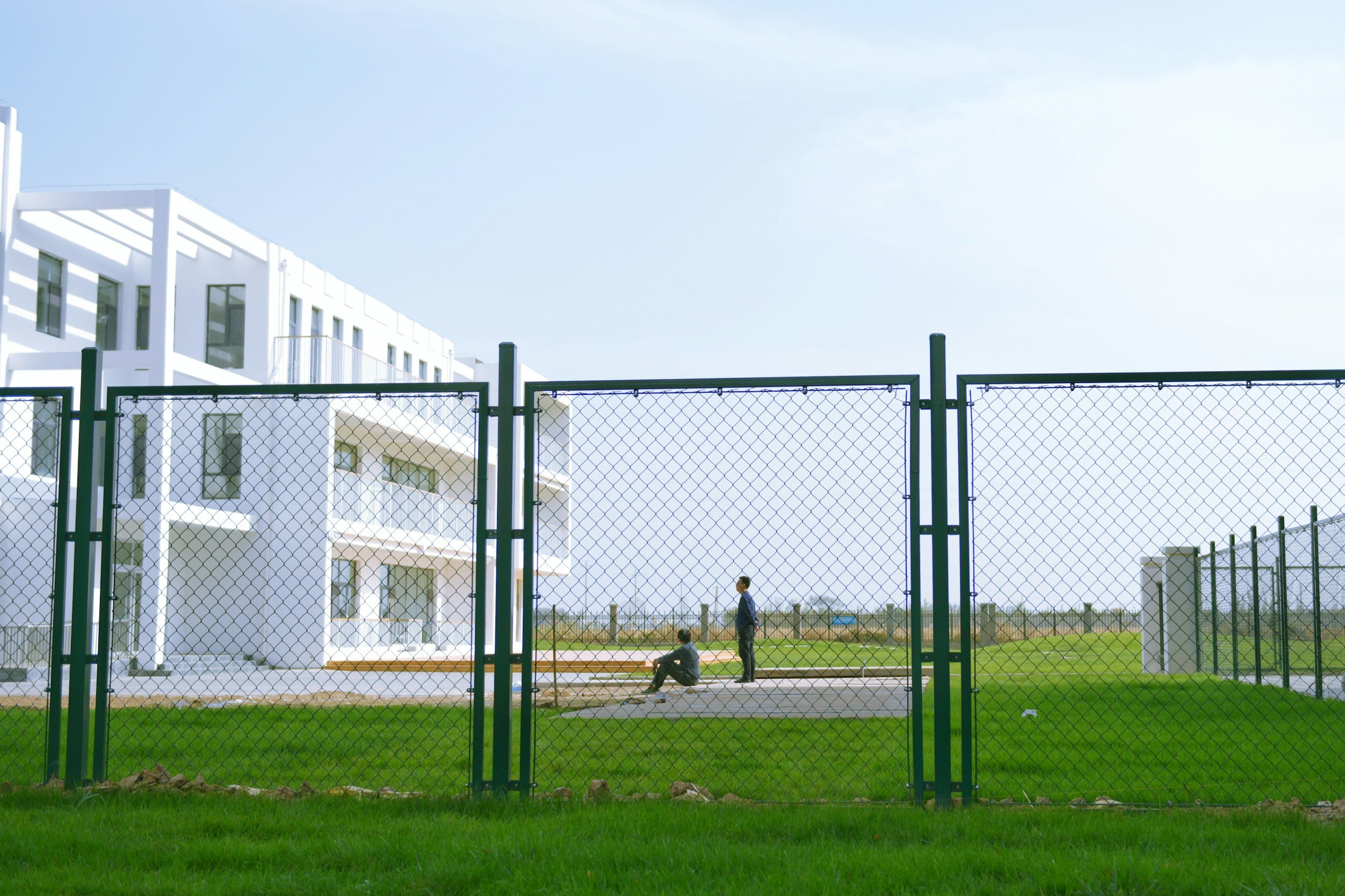 Two people sit on a concrete surface outside a modern white apartment building, behind a tall green chain-link fence under a clear blue sky.