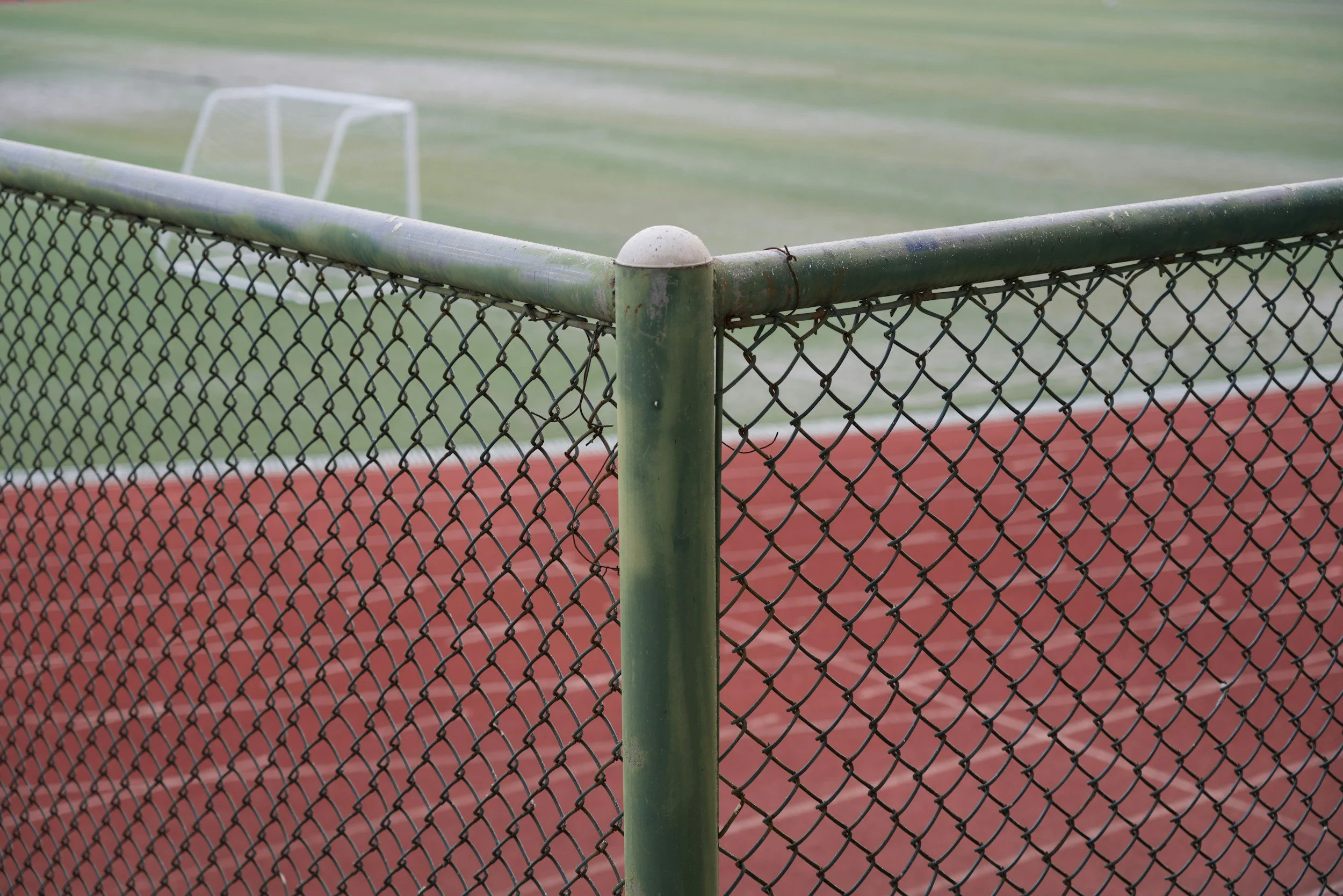 A close-up view of a chain-link fence surrounding a sports field, with the focus on the corner post and a blurred background of a green court and a small goal post.