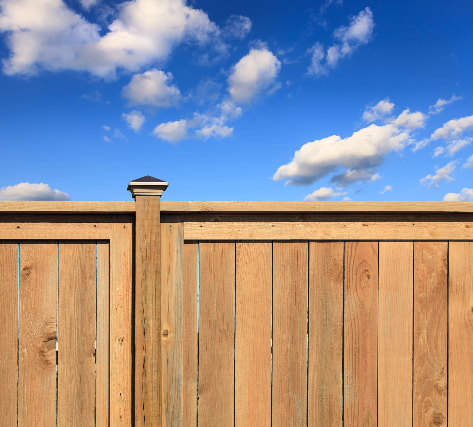 A wooden fence with vertical planks and a top horizontal rail, set against a blue sky with white clouds.