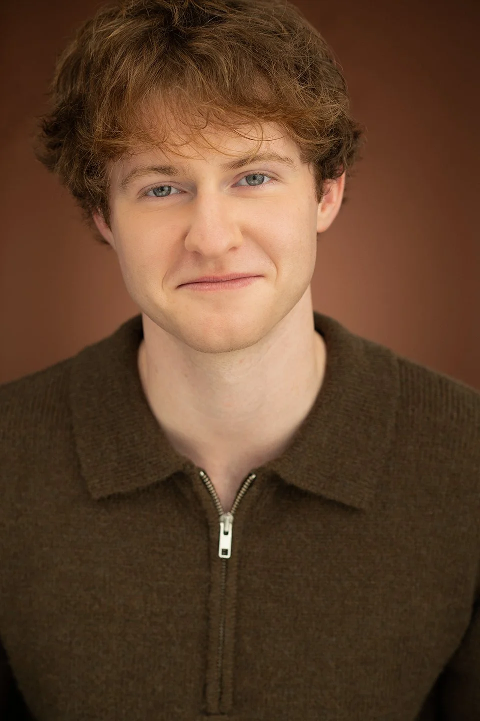Head-and-shoulders portrait of a young man with curly auburn hair and blue eyes, wearing a brown zip-up sweater, smiling softly against a brown background.