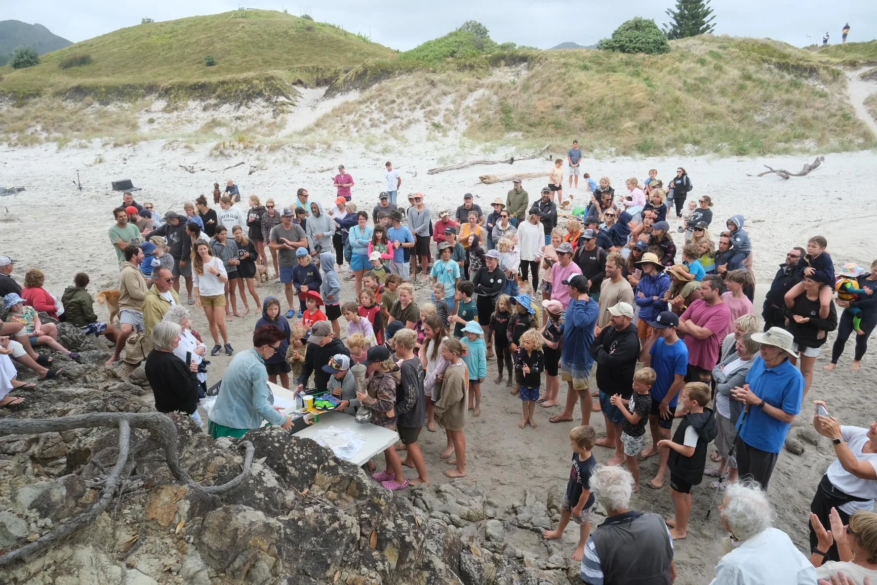 A group of people gathered on a beach for an outdoor event, with some sitting on rocks and others standing in line or watching. There are hills and trees in the background, and the scene appears to be a community gathering or activity.