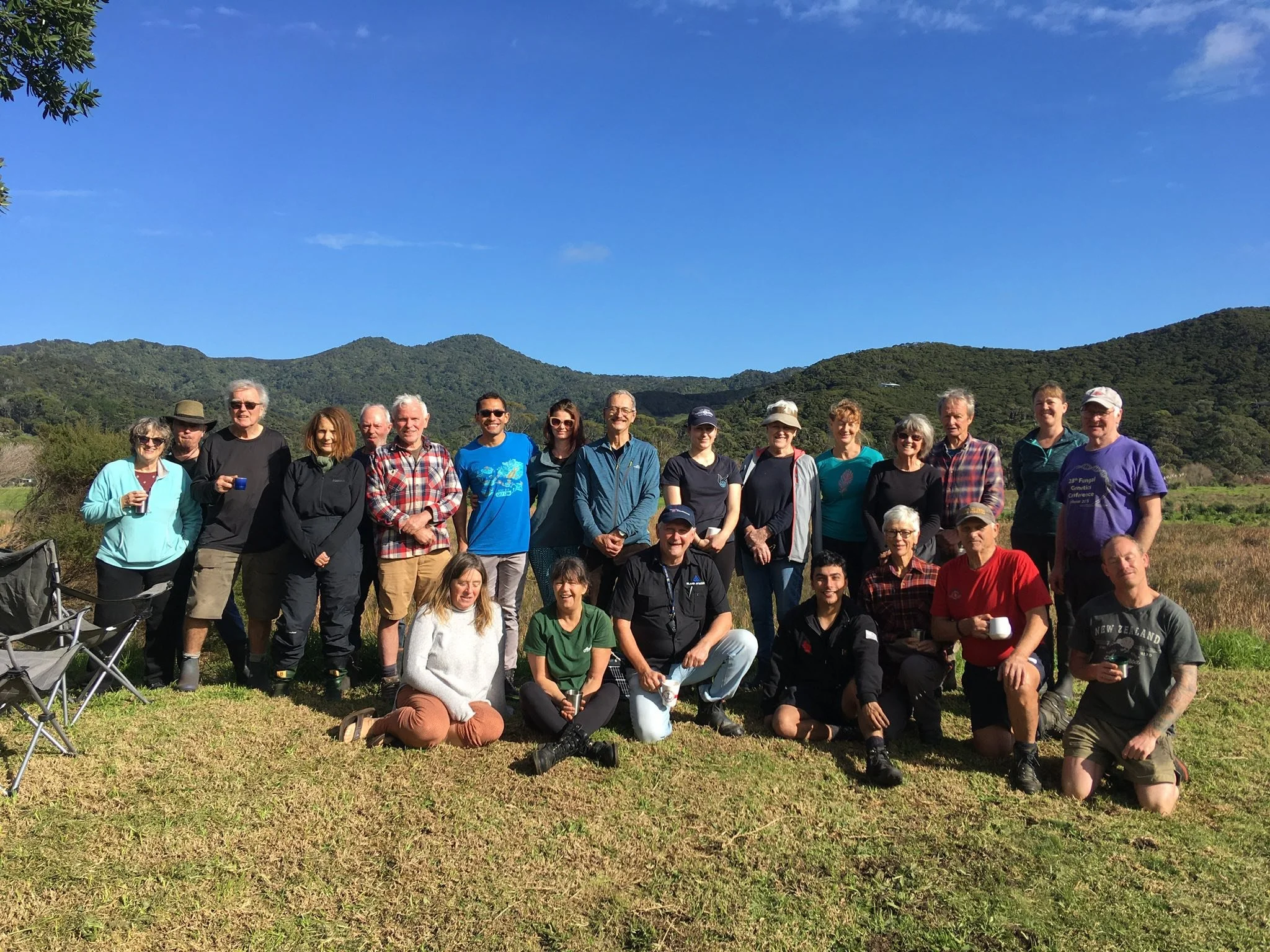 A group of 21 people outdoors in front of green hills and a clear blue sky, some holding cups or cameras, posing for a group photo on a grassy area.