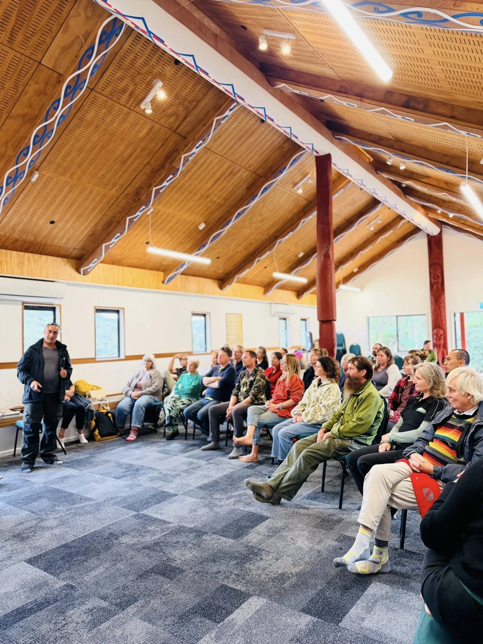 A group of people attending a presentation in a room with a wooden ceiling, some sitting and some standing, with windows in the background.