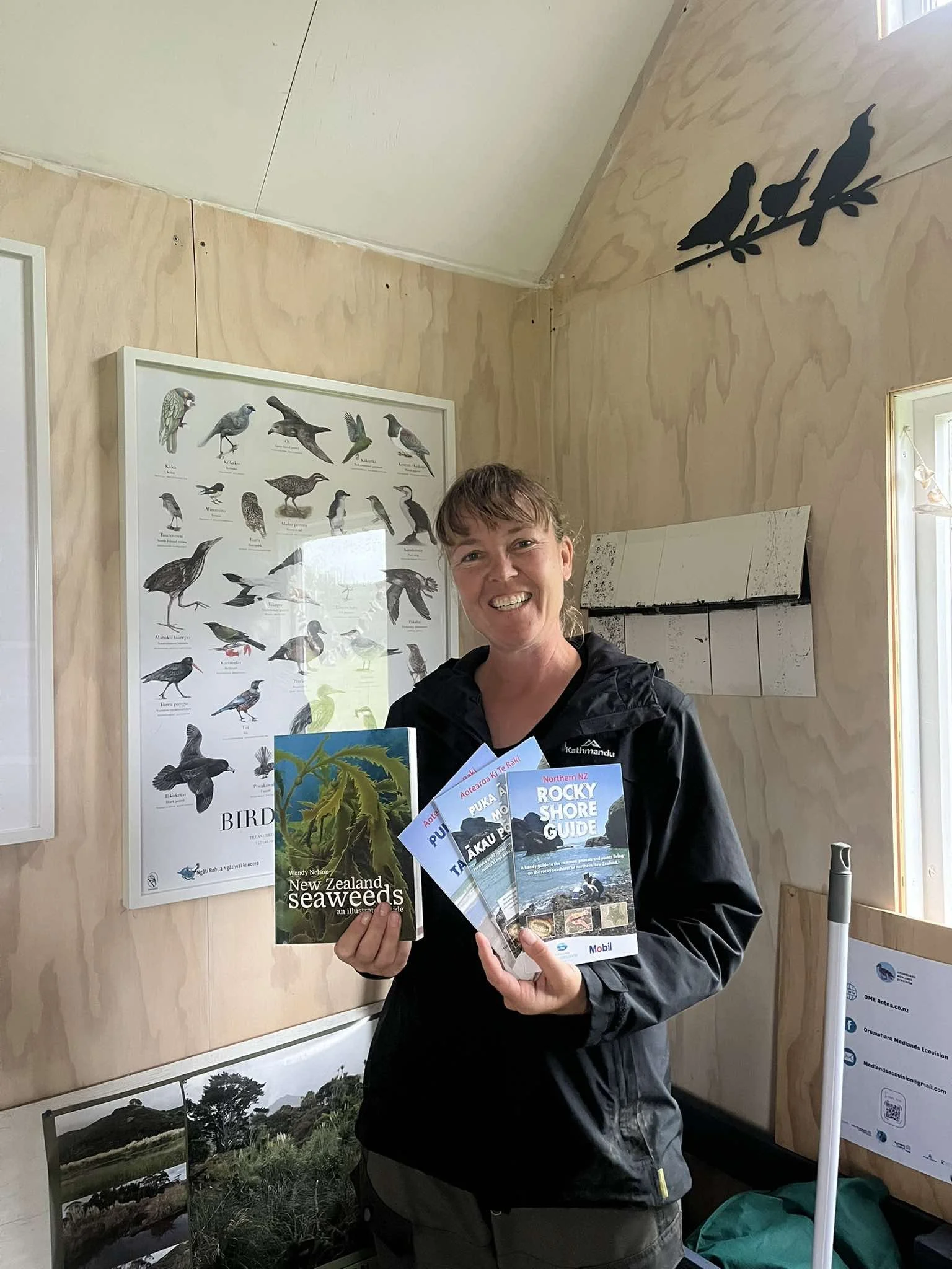 A woman smiling and holding three brochures about New Zealand seaweeds, the Auk rainforest, and the Rocky Shore Guide, standing inside a room decorated with bird posters and silhouettes, with some landscape photographs on a table.