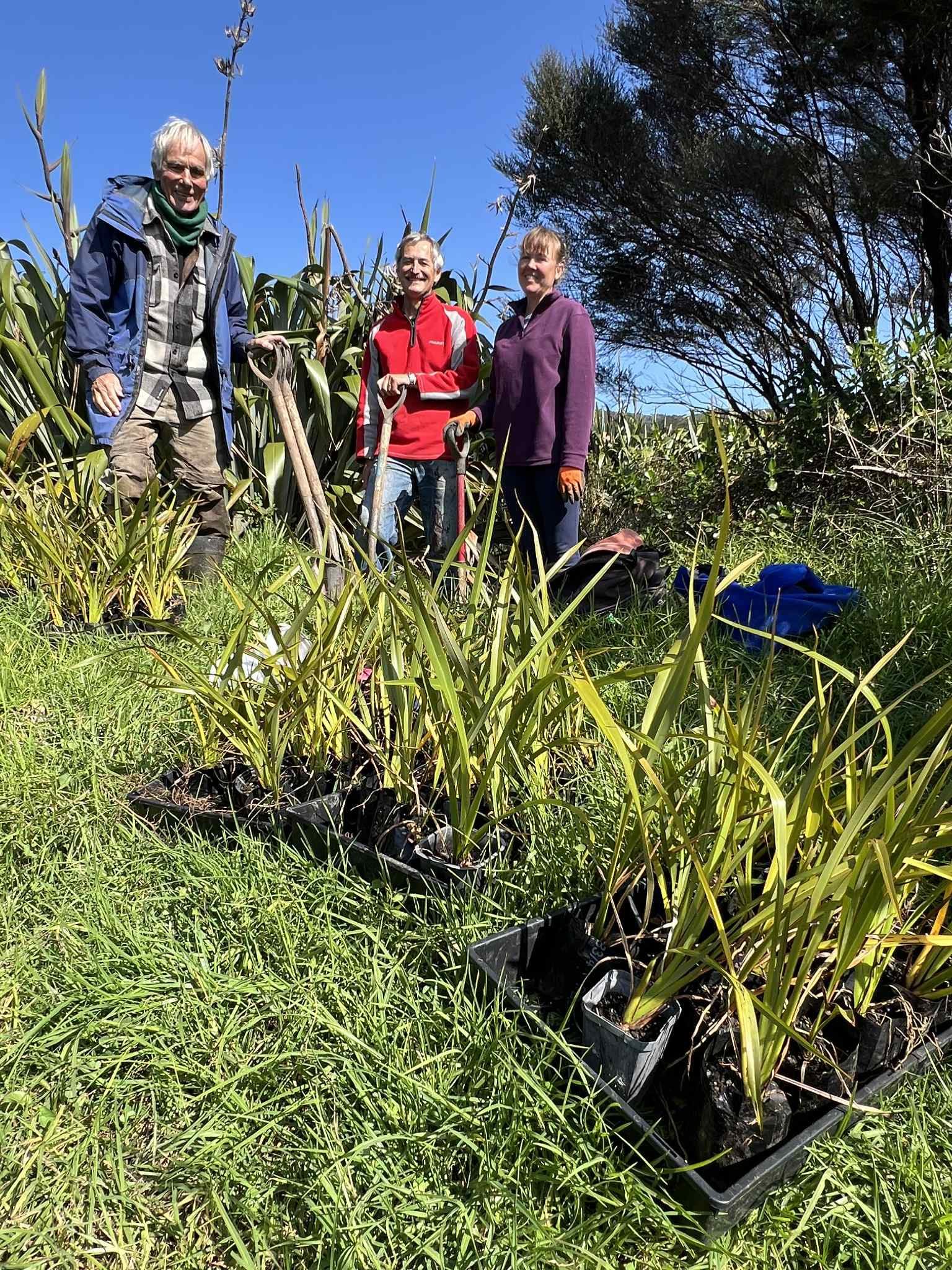 Three people standing outdoors in a grassy field with plants and trees, smiling at the camera on a sunny day.
