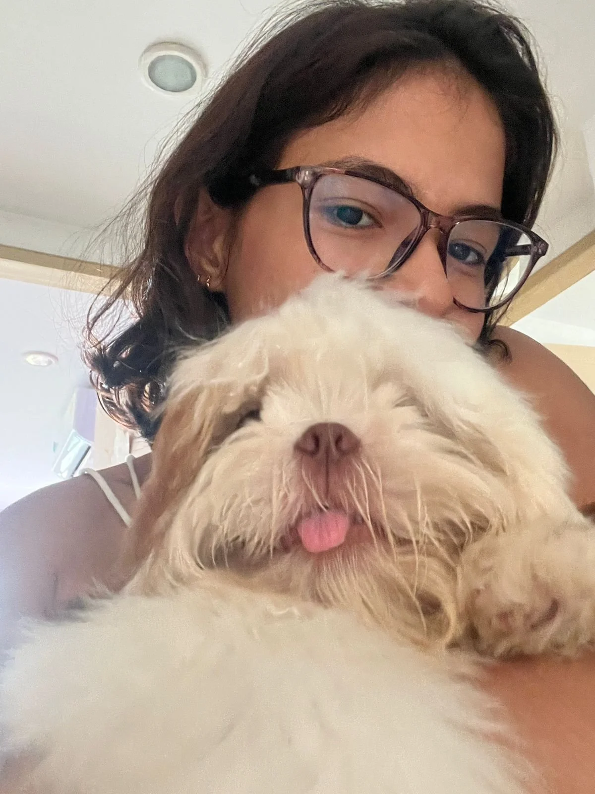 A woman with glasses and dark hair taking a close-up selfie with her fluffy white dog, who is sticking out its tongue.
