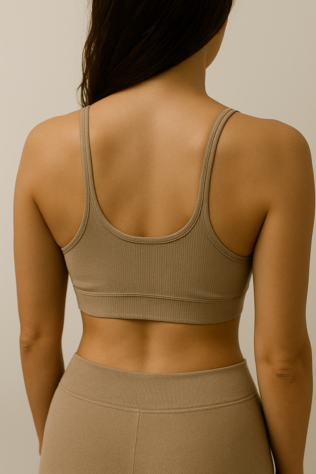 A woman with long, dark hair wearing a tan athletic crop top and matching high-waisted leggings, facing away from the camera in a neutral setting.