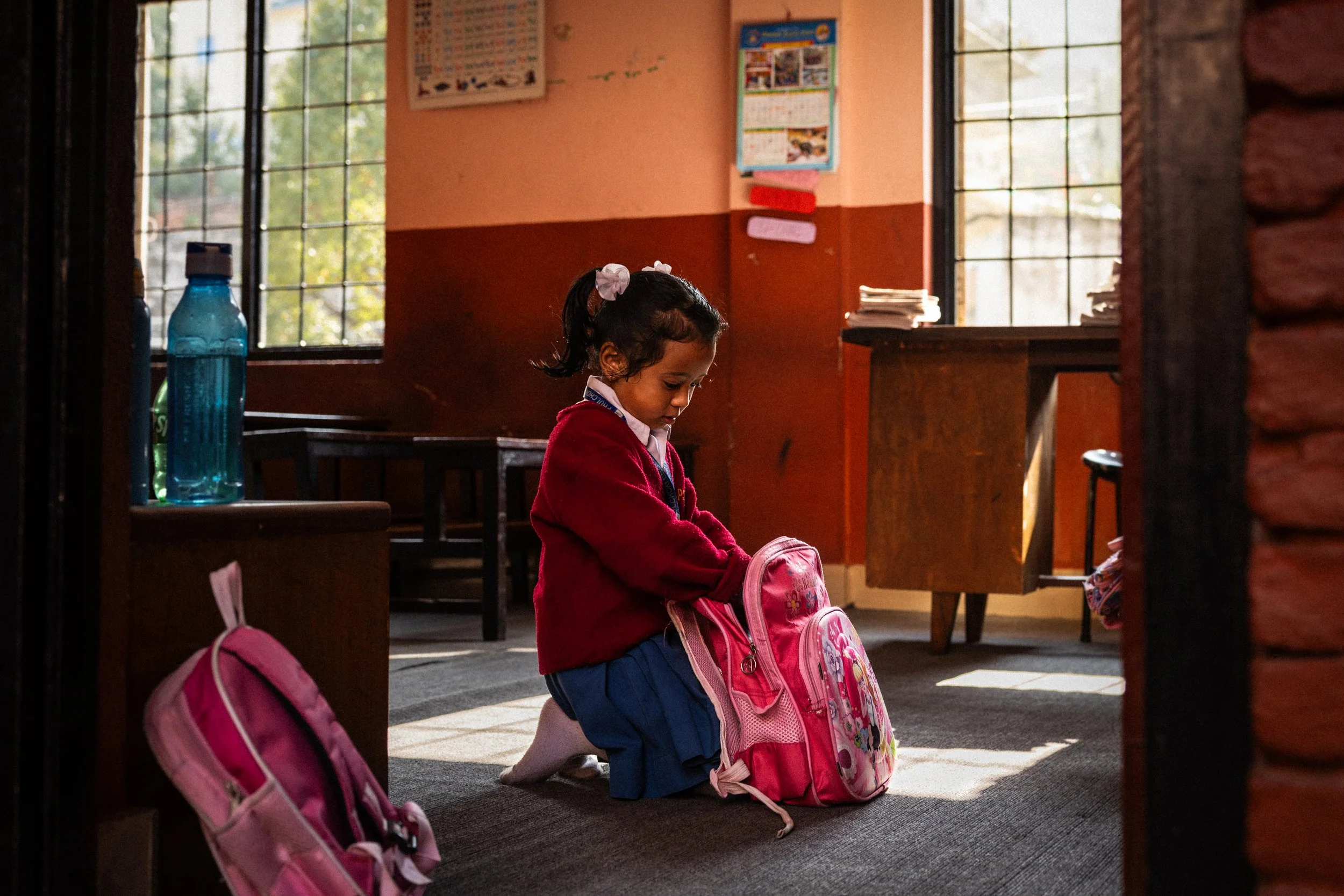 Una bambina che si mette lo zaino rosa in una scuola con pareti arancioni e grandi finestre, seduta sul pavimento mentre si prepara per la scuola.