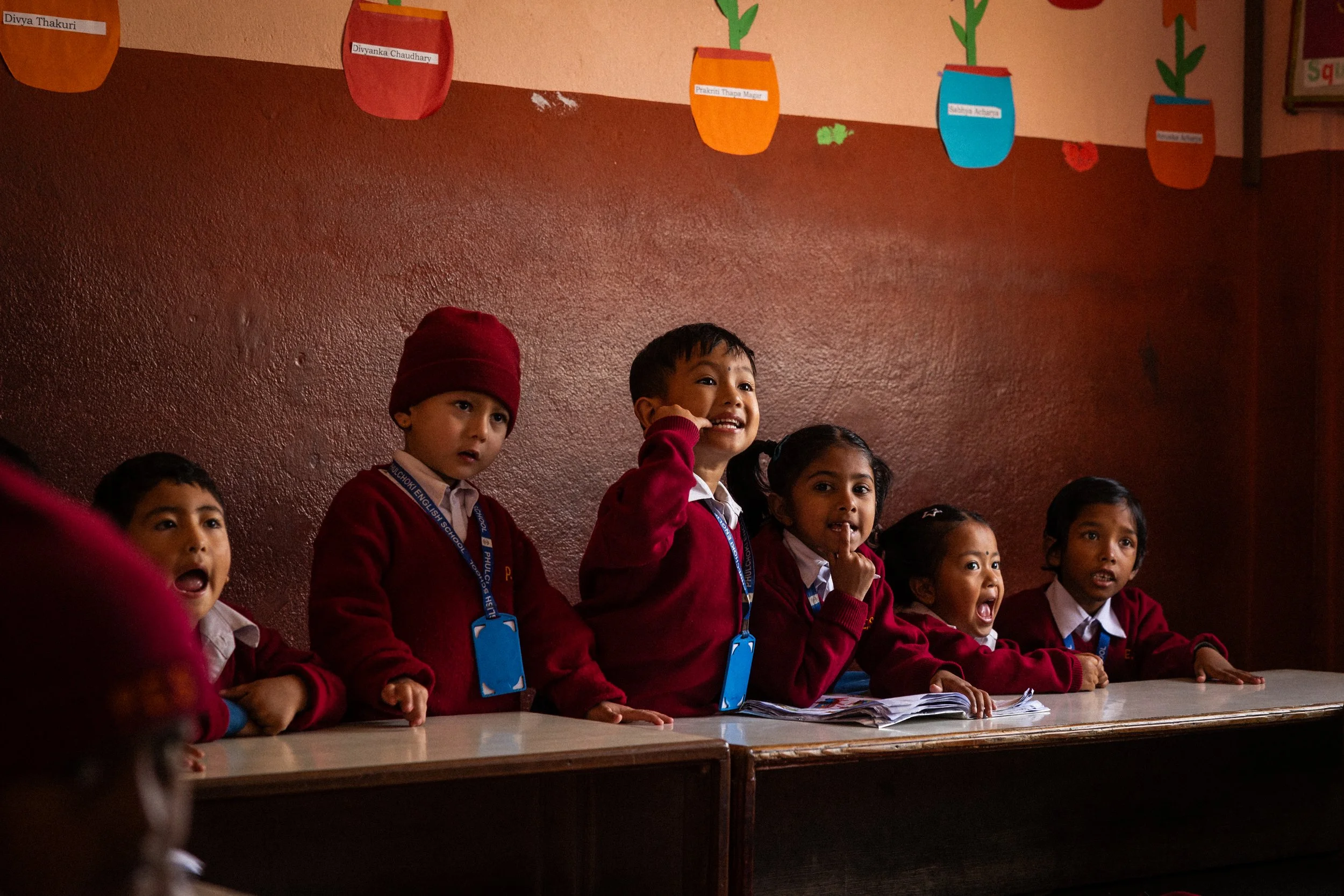 Gruppo di bambini in uniforme scolastica seduti dietro un banco, alcuni che sorridono o fanno espressioni di stupore, in una classe con pareti rosse e decorazioni colorate sopra il banco.