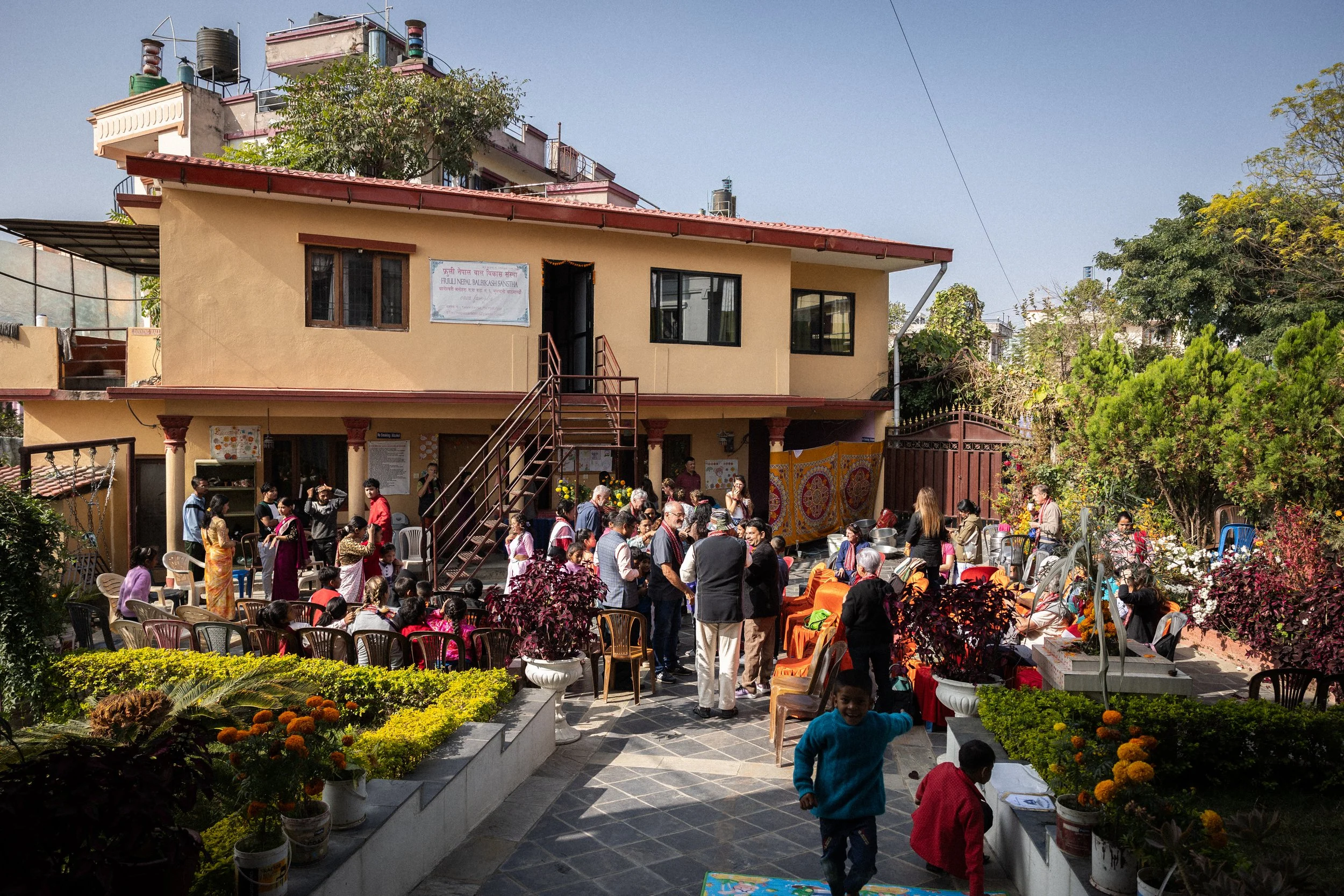 Un cortile affollato con molte persone partecipanti a un evento o riunione, alcune sedute, altre in piedi, con un edificio a due piani sullo sfondo, circondato da piante e fiori colorati.