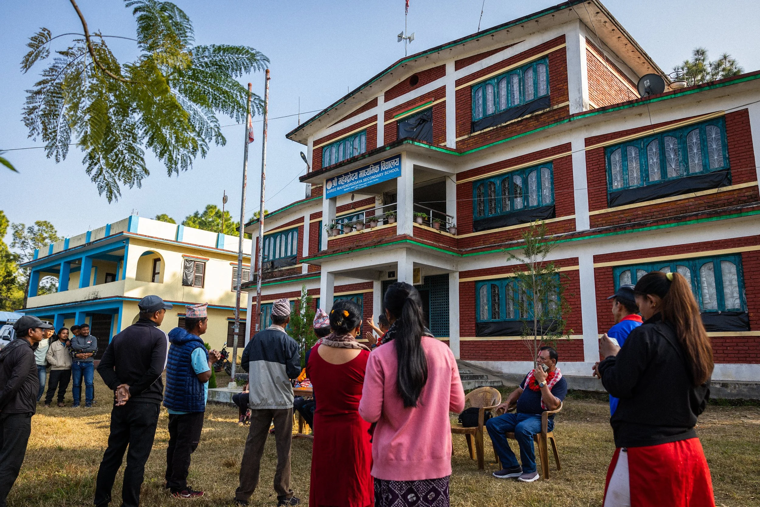 Gruppo di persone che si riuniscono all'esterno di un edificio scolastico, alcuni siedono e altri stanno in piedi, durante un evento o una celebrazione.
