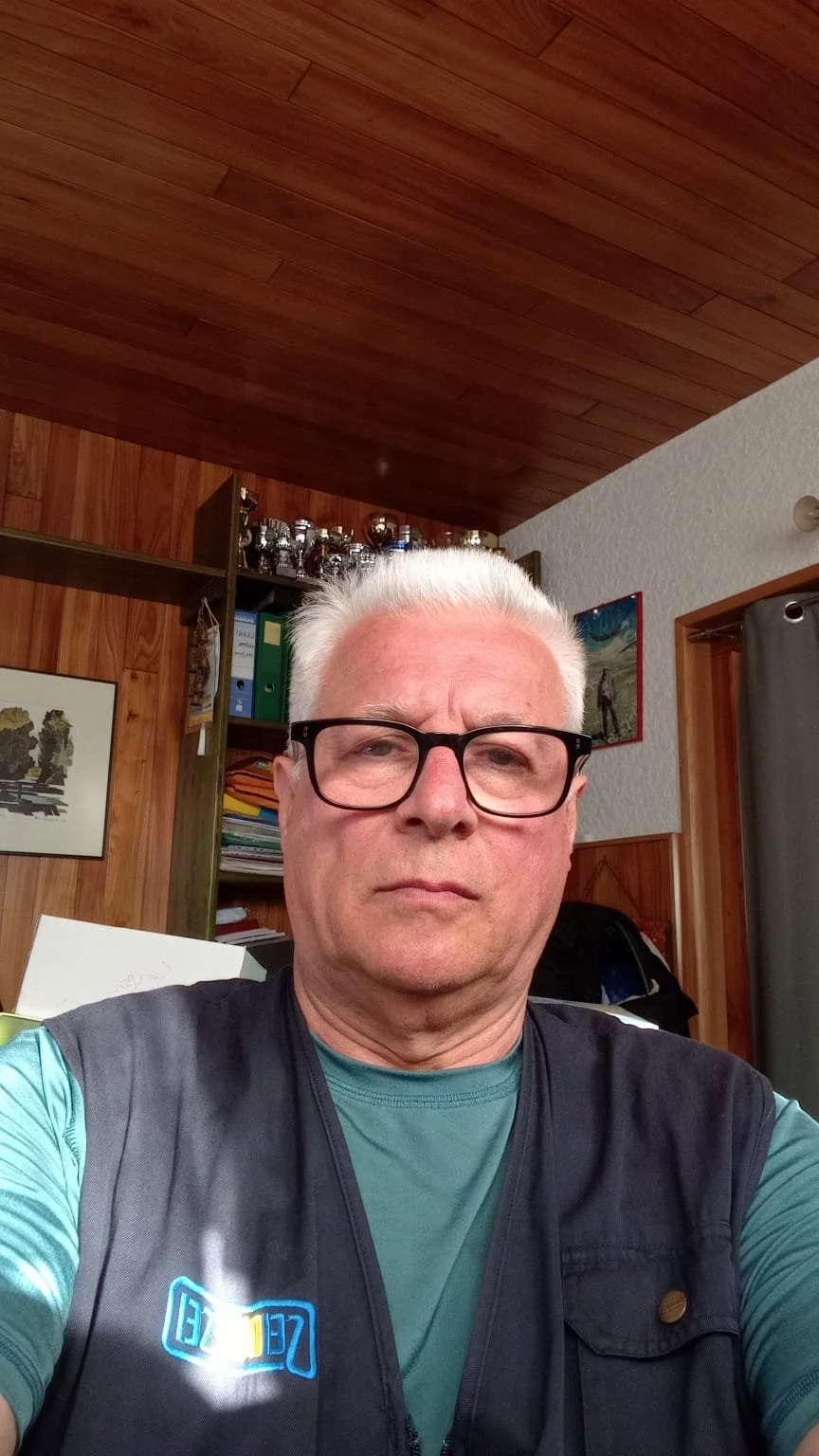Uomo con occhiali scuri e camicia verde in una stanza con pareti di legno e oggetti decorativi sullo sfondo.
