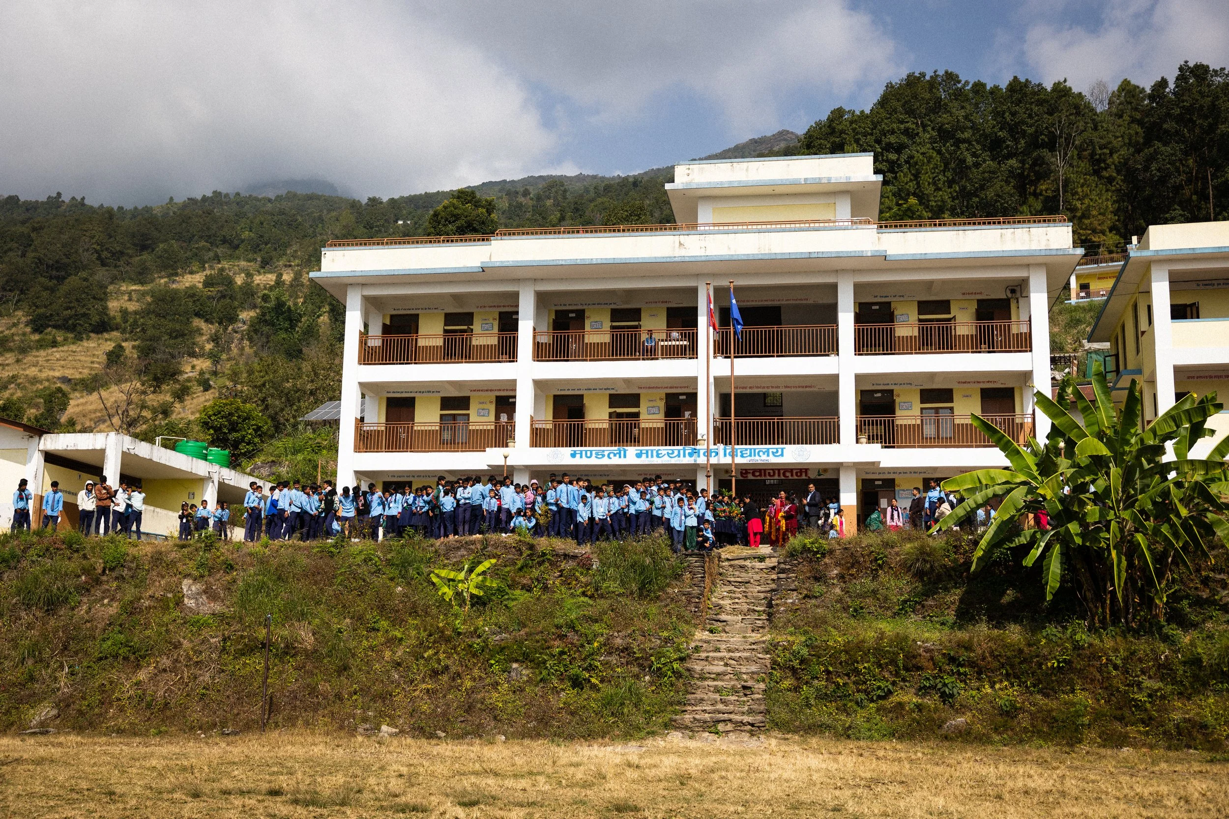 Scuola elementare con molti studenti in uniforme blu fuori dall'edificio, situata in una zona montuosa, con bandiere davanti.