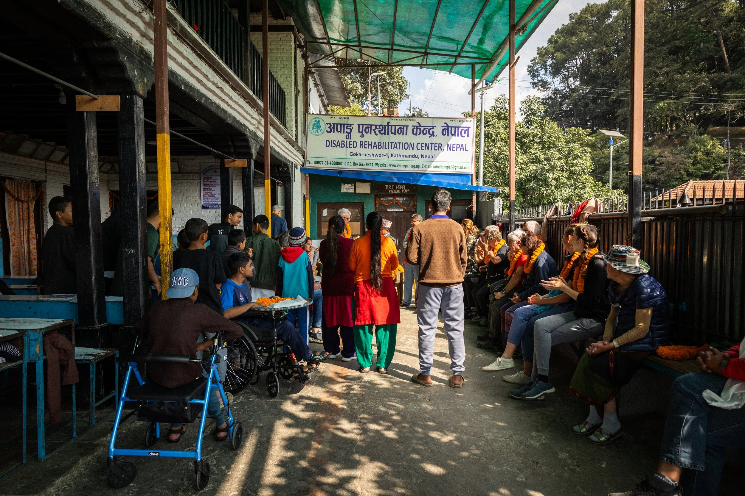 Persone sedute e in piedi davanti a un centro di riabilitazione disabile a Kathmandu, Nepal, con alcuni individui che indossano collane di marigold, sotto un'insegna gialla e bianca.