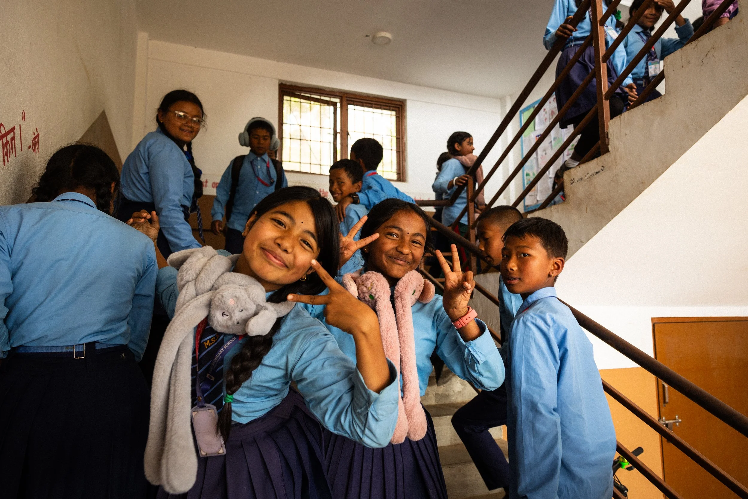 Gruppo di studenti sorridenti in uniforme scolastica in un edificio con scale, alcuni fanno segni di pace con le dita.
