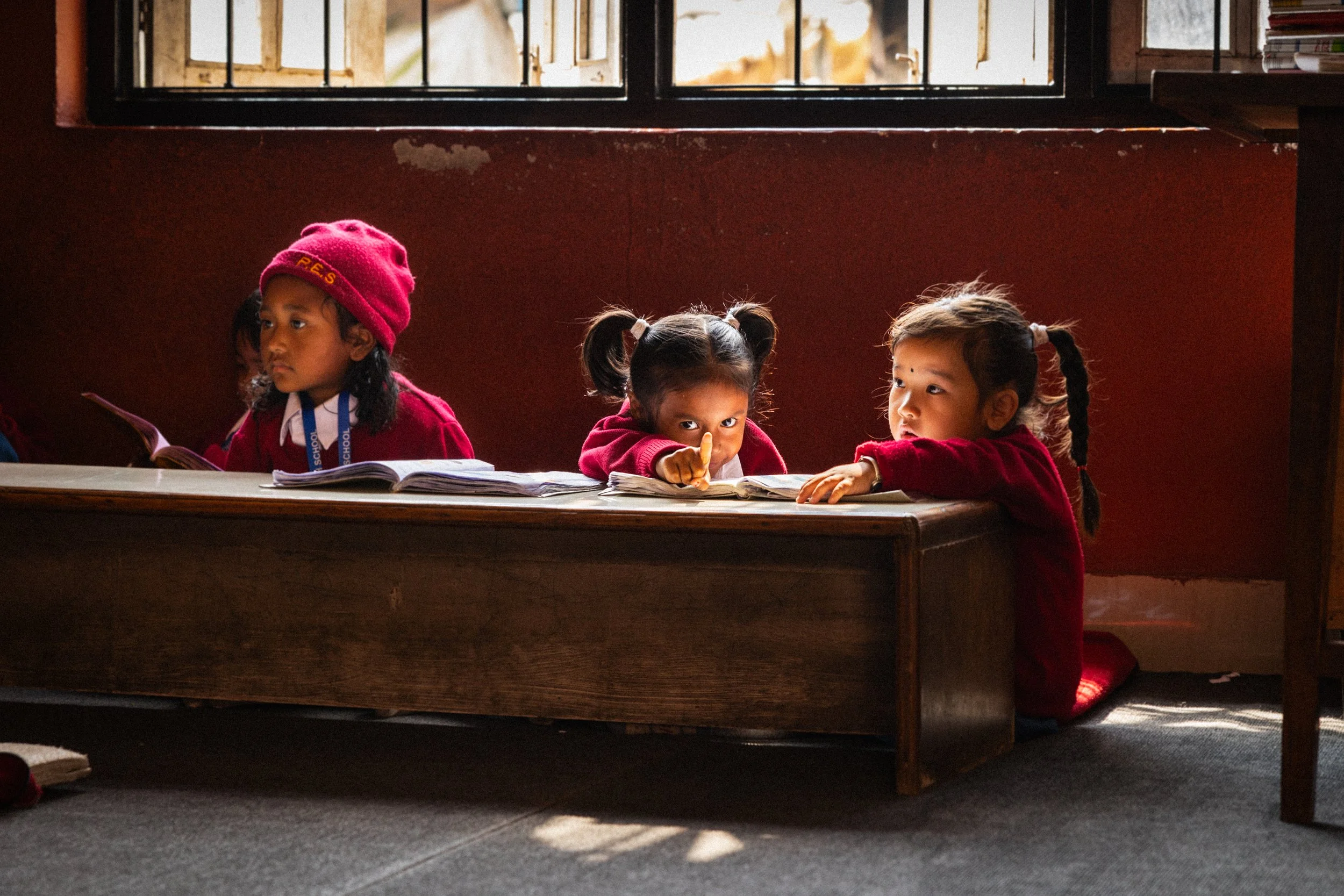 Tre bambine sedute a terra vicino a un banco scolastico, in una classe con pareti rosse e una finestra aperta dietro.
