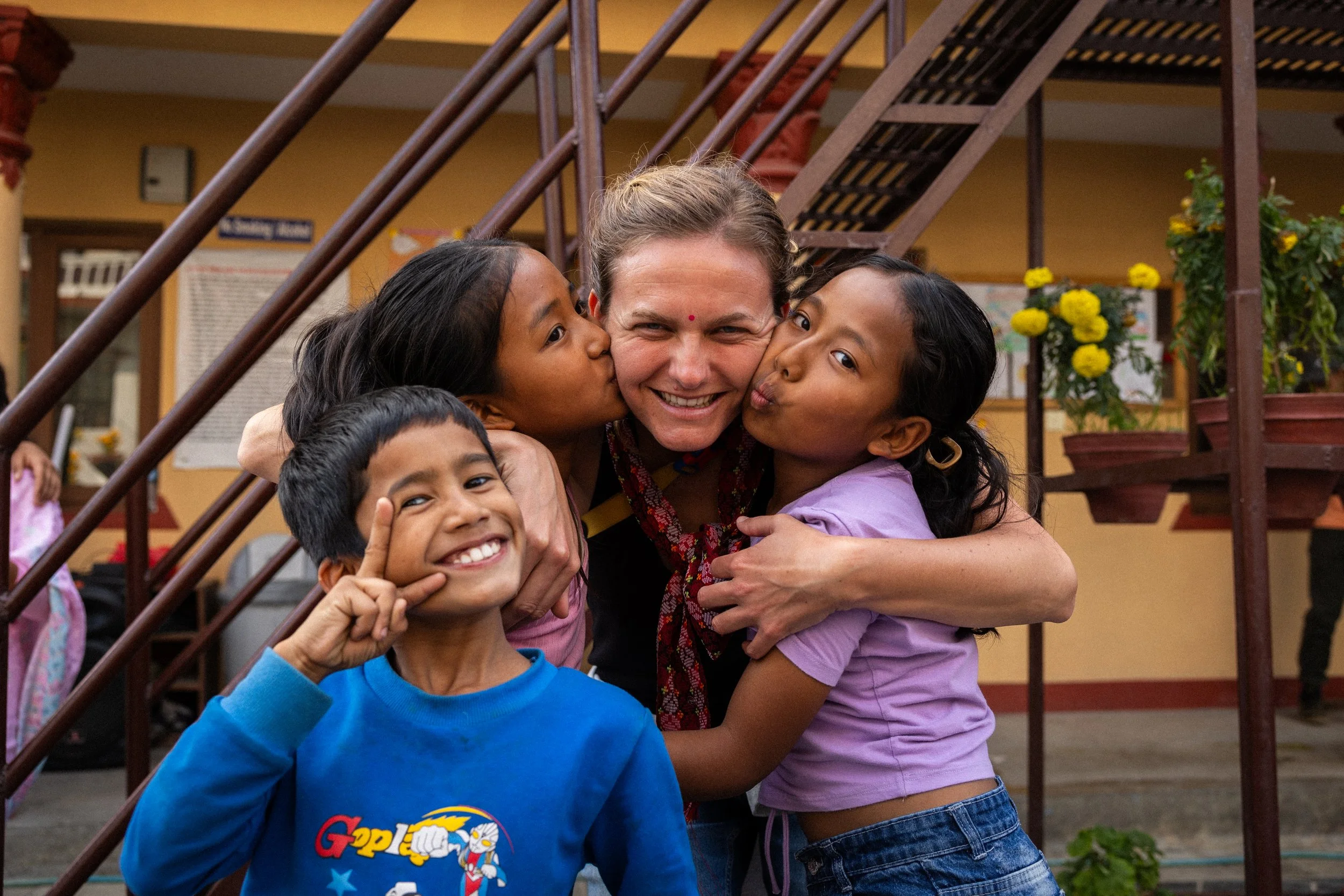 Donna circondata da tre bambini, che le dà baci e abbracci, all'aperto con una scala in metallo e fiori gialli sullo sfondo.