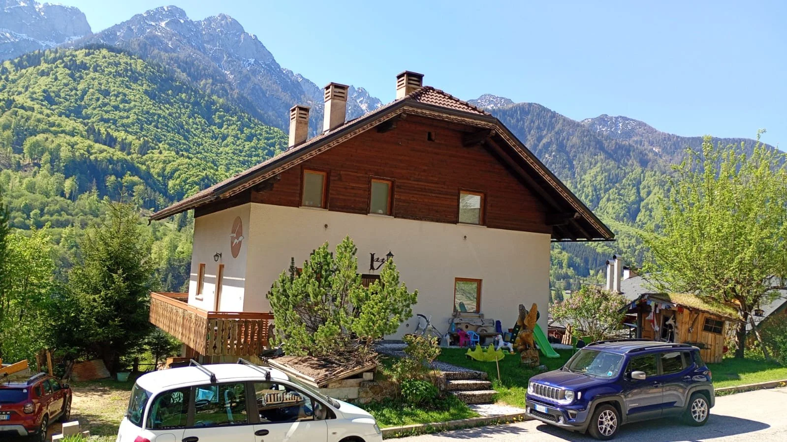 Casa di montagna con giardino, auto e montagne sullo sfondo.