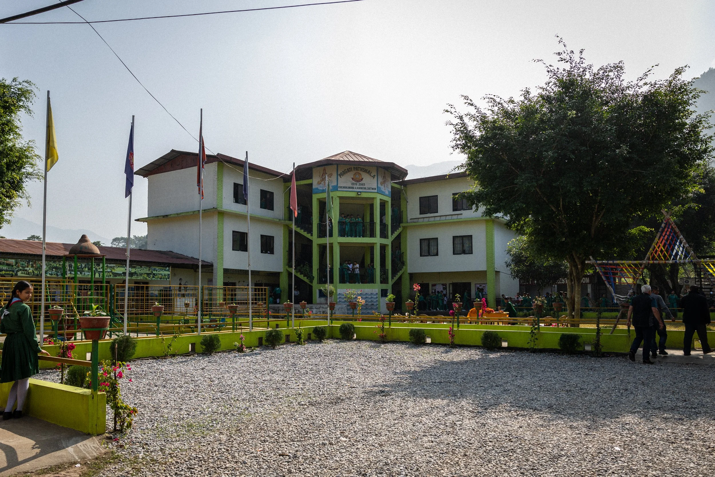 Scuola in stile edificio di campagna con facciata bianca e dettaglio verde, giardino con aiuola e panchine, persone che camminano e studenti in uniforme. Al centro, due pianianni con un portico e bandiere che sventolano.