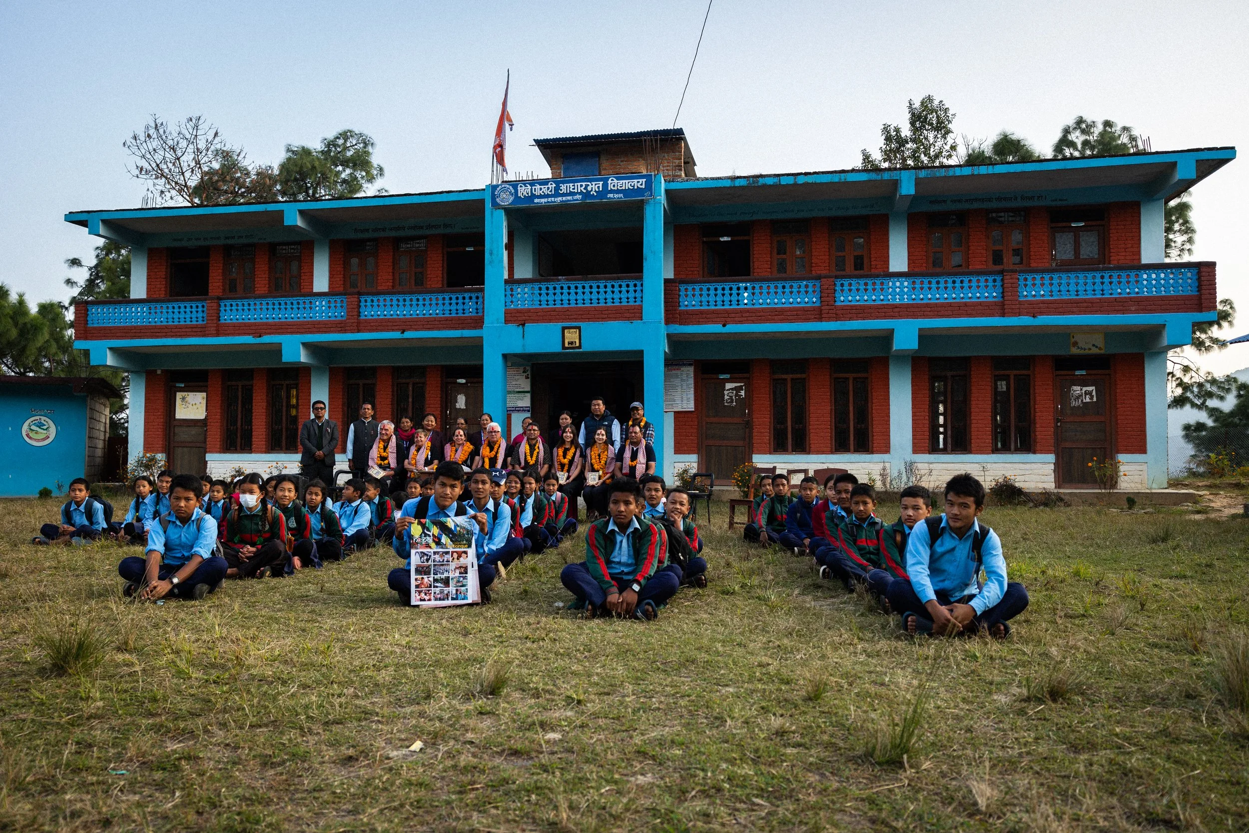 Gruppo di studenti seduti in primo piano davanti a una scuola, con alcuni insegnanti e funzionari alle spalle, in un'area all'aperto, in un paesaggio con alberi e un edificio scolastico colorato.