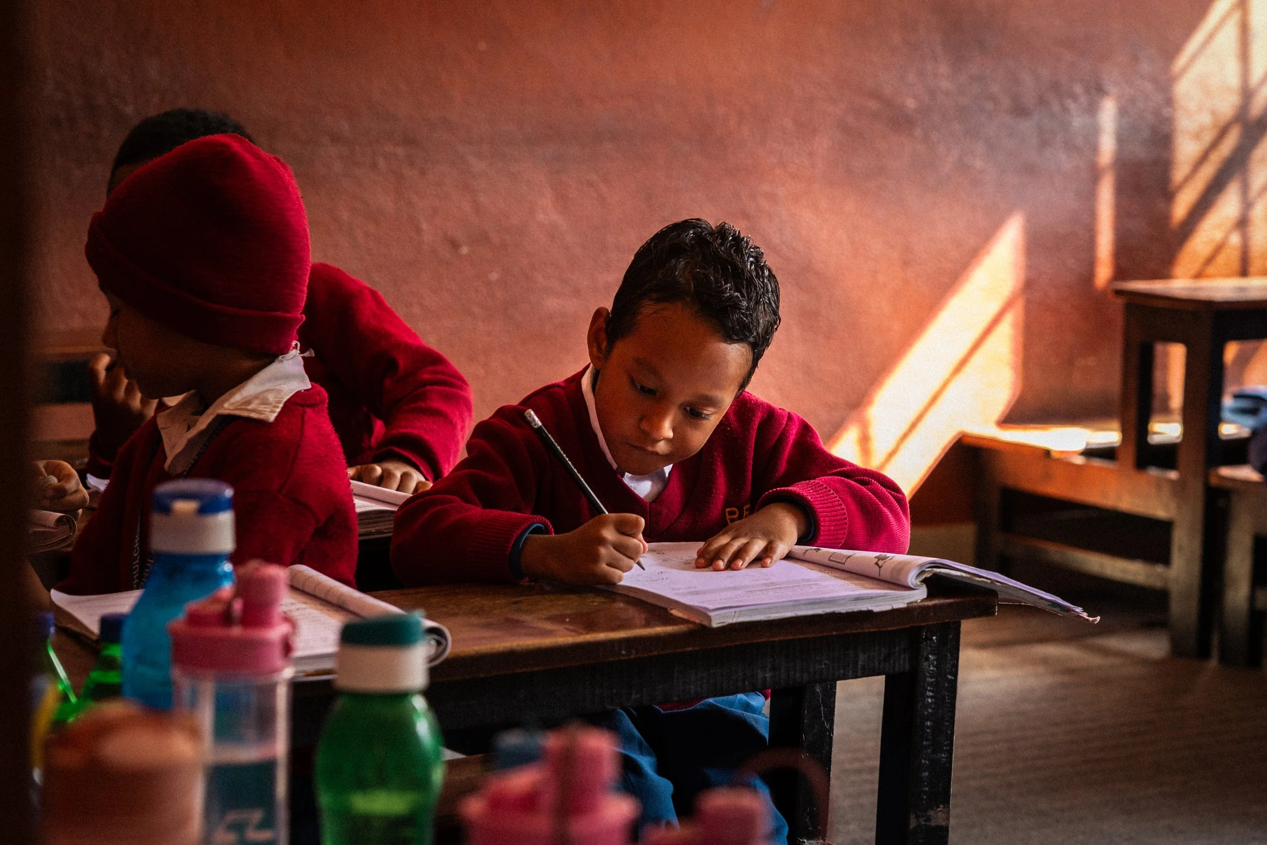 Bambini in classe che studiando, uno scrive e l'altro si dedica ai compiti, con bottiglie di acqua sul banco e una parete di colore rosso.