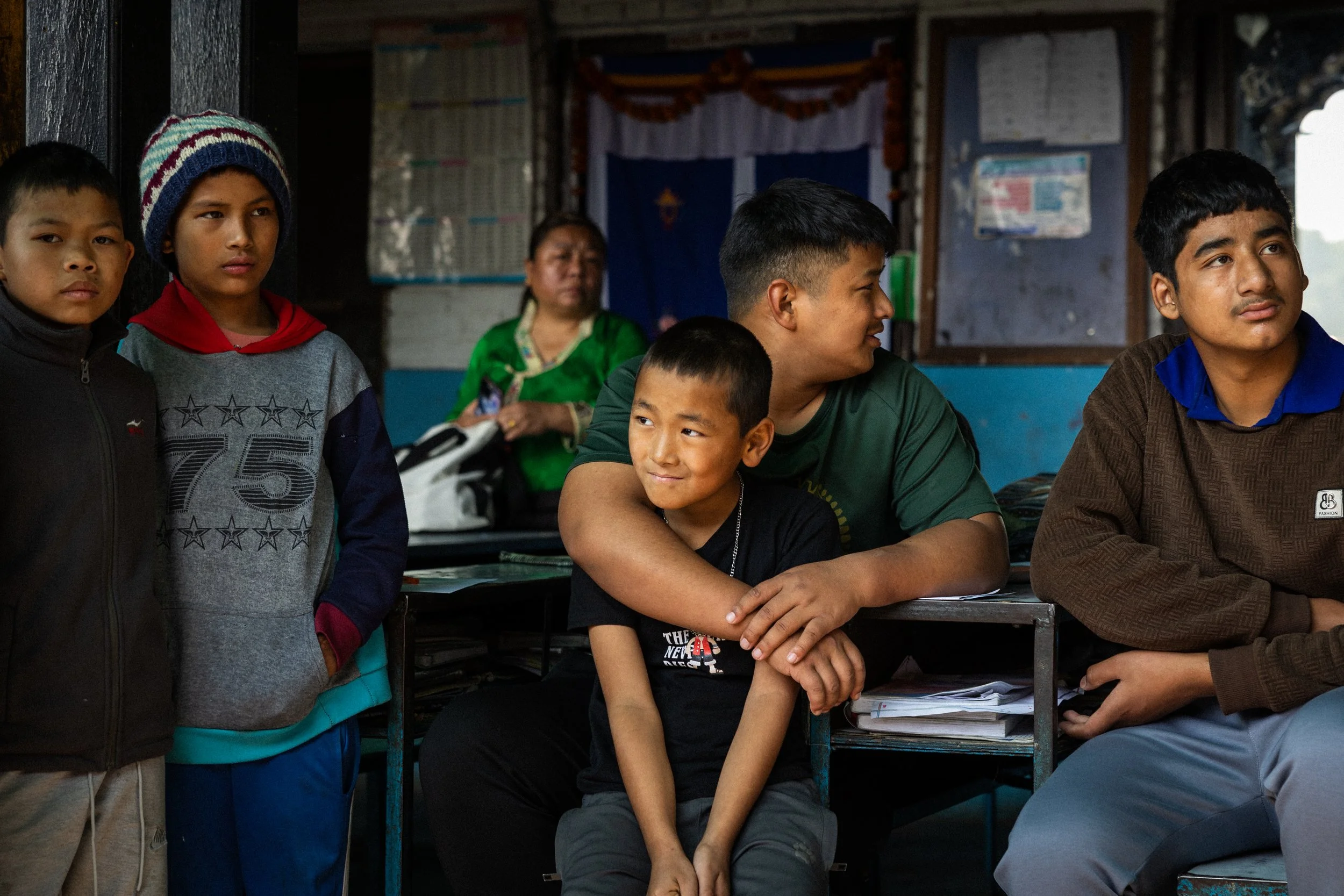 Gruppo di bambini e adolescenti seduti e in piedi in una stanza scolastica, alcuni che osservano avanti, con sfondo di bacheca e pareti blu, qualcuno con abbigliamento casual e cappello colorato.