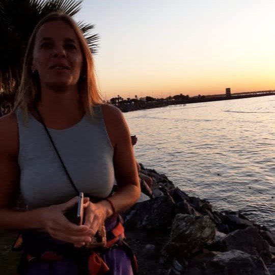 Donna con abbigliamento estivo in riva a un fiume al tramonto, con palme sullo sfondo.