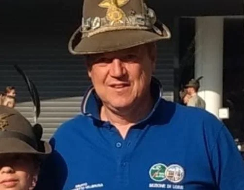 Uomo che indossa un cappello da esploratore con decorazioni e una maglietta blu, con altri persone in uno sfondo esterno