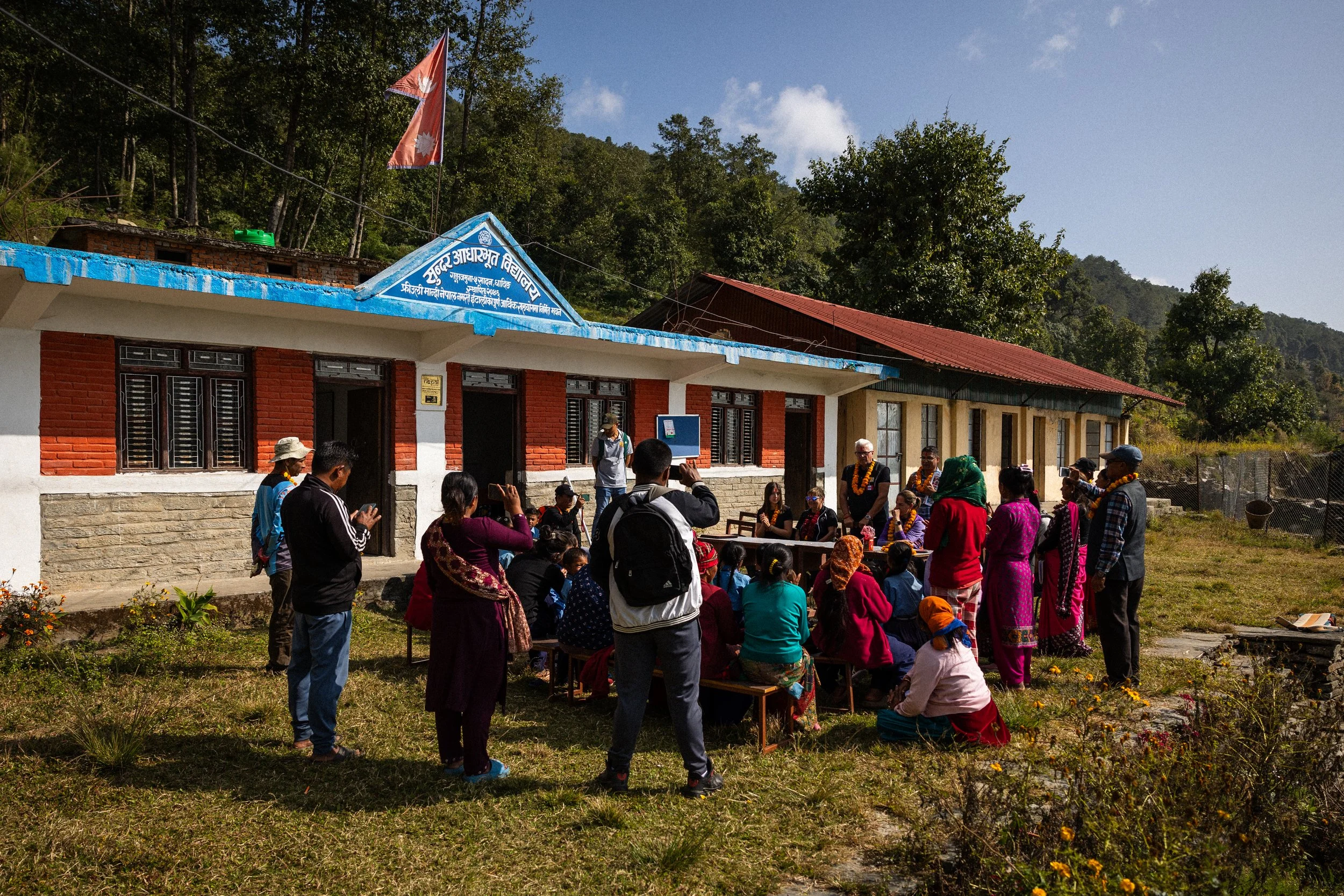 Gruppo di persone di fronte a un edificio con una bandiera nepalese, apparentemente per una cerimonia o un evento culturale, con clima soleggiato e un paesaggio di colline e alberi sullo sfondo.