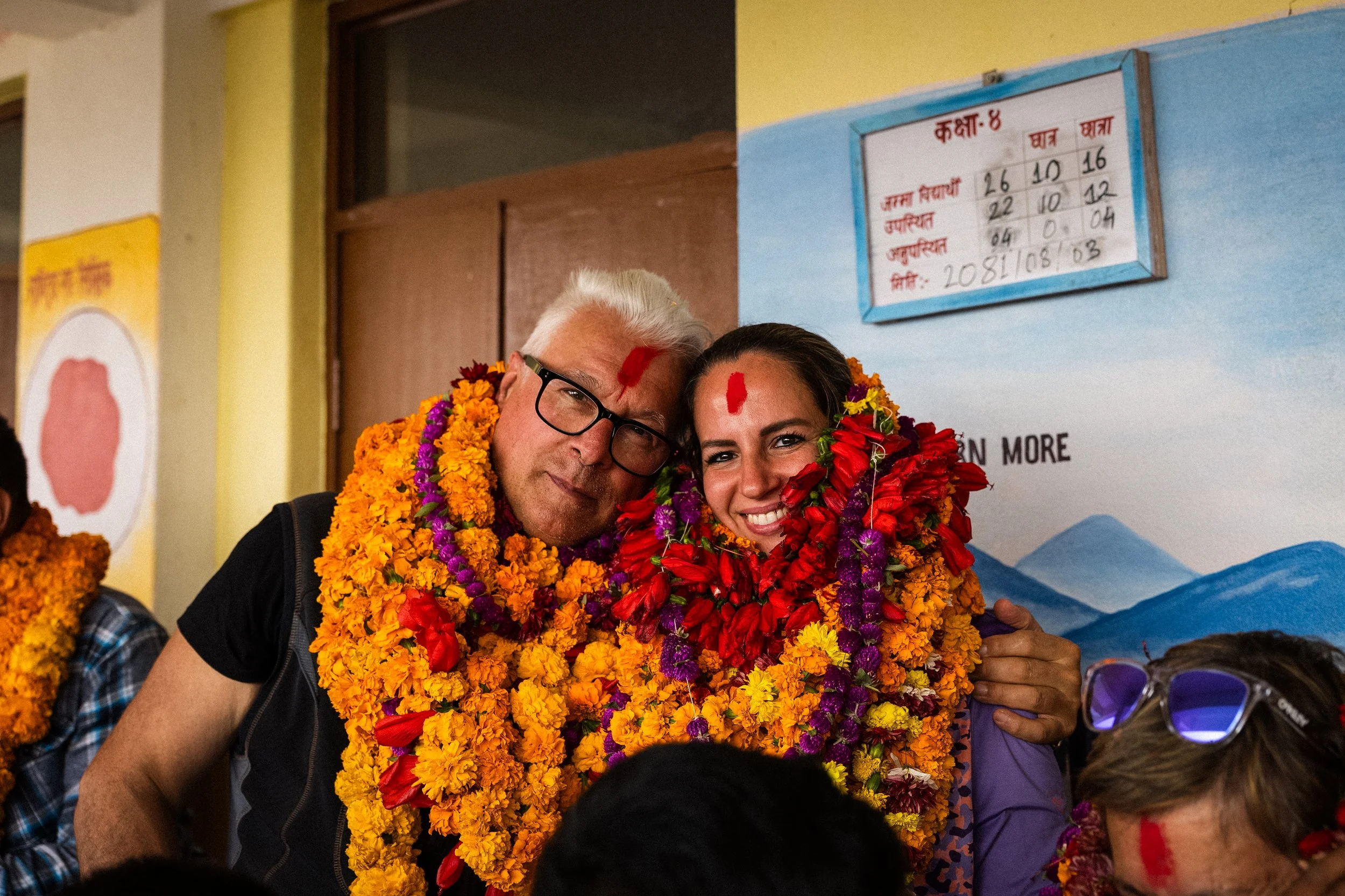 Due persone, un uomo con capelli bianchi, occhiali e una donna con capelli scuri, sorridenti, indossano molte collane di fiori di diverse colori, celebrando un evento, con pareti decorate e segnaletica in lingua nepalesi sullo sfondo.