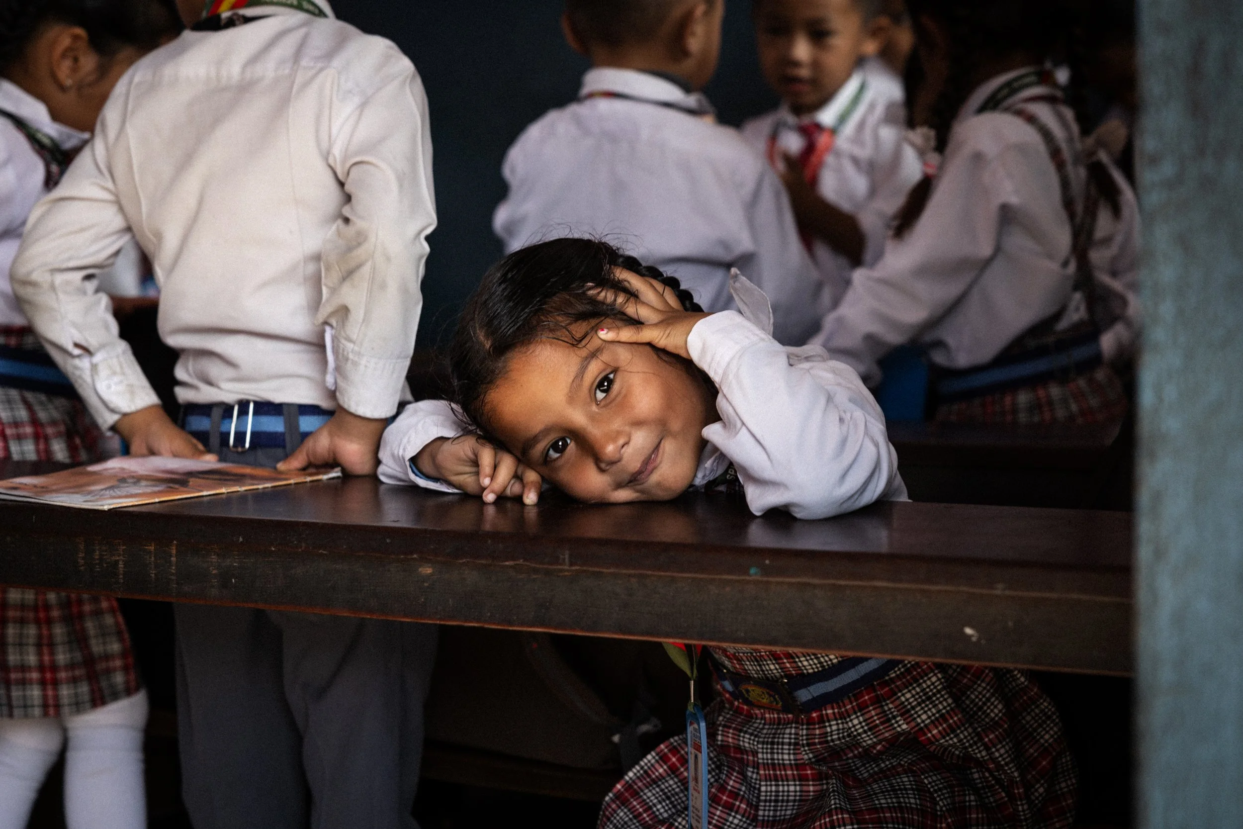 Una ragazza indossa una camicia bianca e una gonna a quadri rossi e neri, si appoggia sul tavolo con la testa appoggiata alle mani, mentre è circondata da altre bambine in uniforme scolastica.