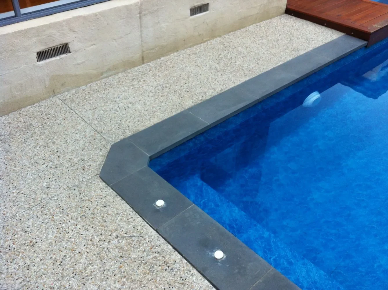 Close-up of a swimming pool with blue water, gray border tiles, and beige and brown surrounding deck.