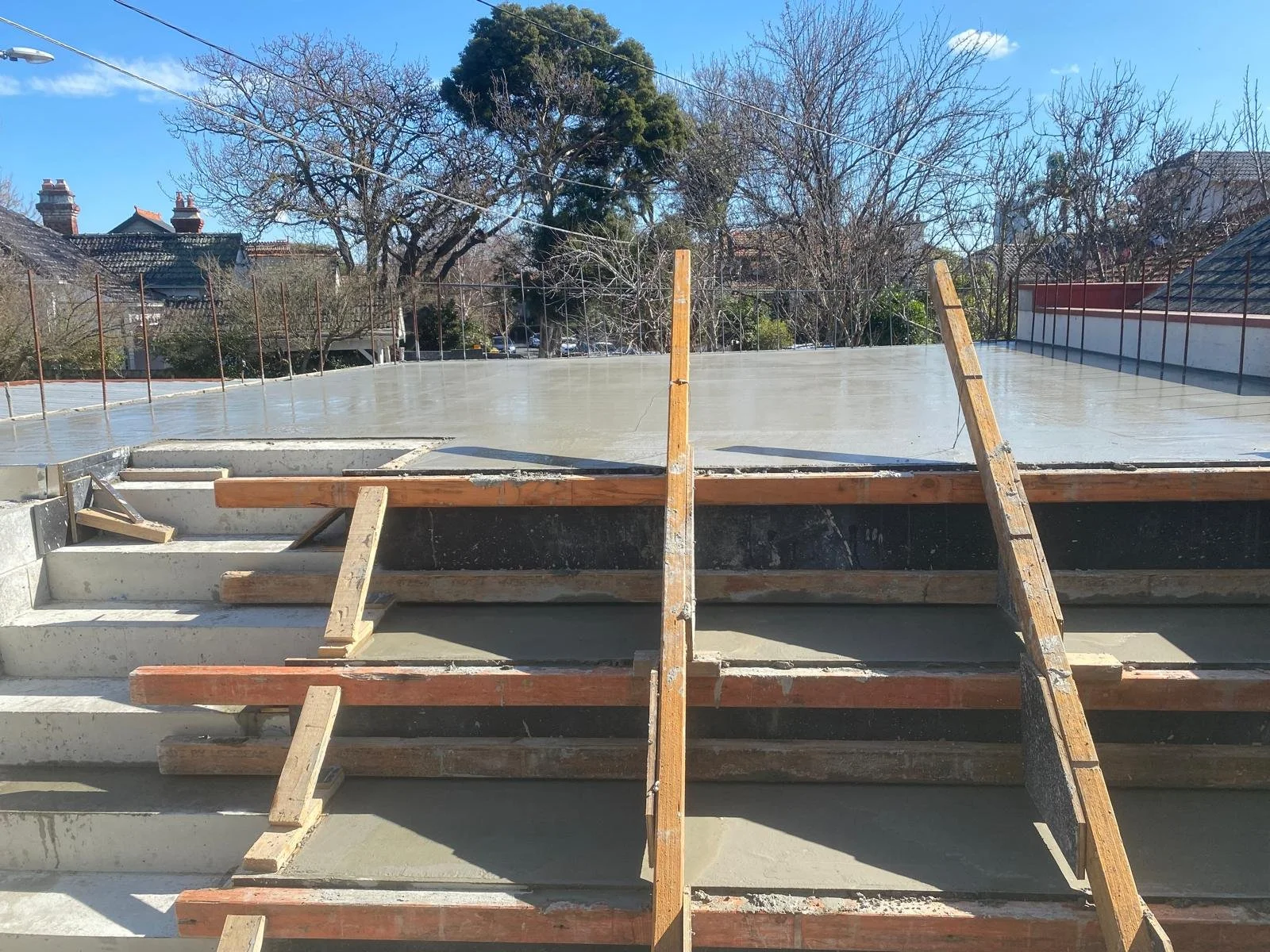 Construction site with concrete stairs and a partially built flat concrete platform or roof, surrounded by wooden scaffolding and ropes, with trees and neighboring houses in the background.
