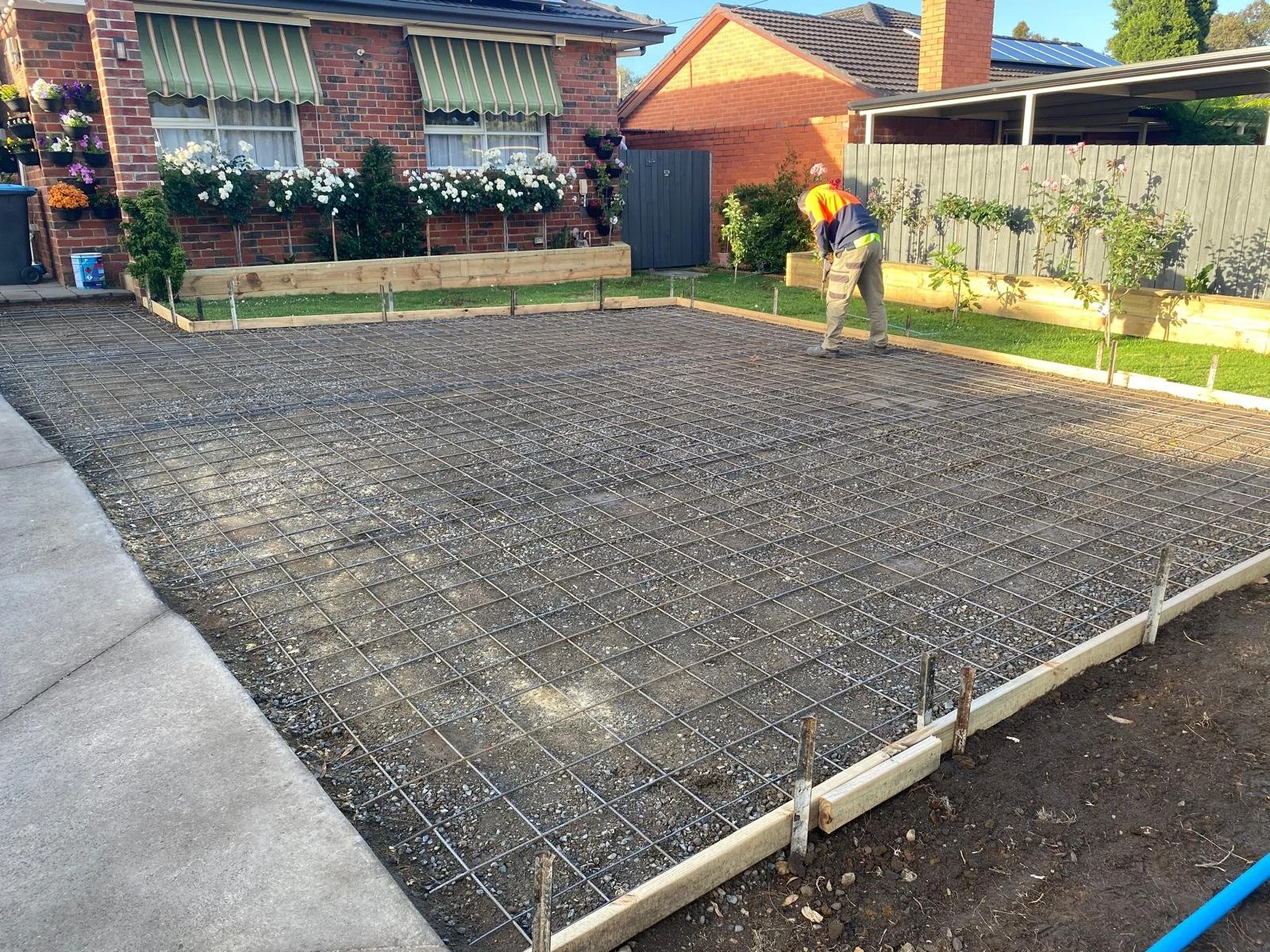 A worker leveling a freshly poured concrete slab in a residential backyard, with a wire mesh grid and wooden formwork in place, surrounded by flower beds and houses.