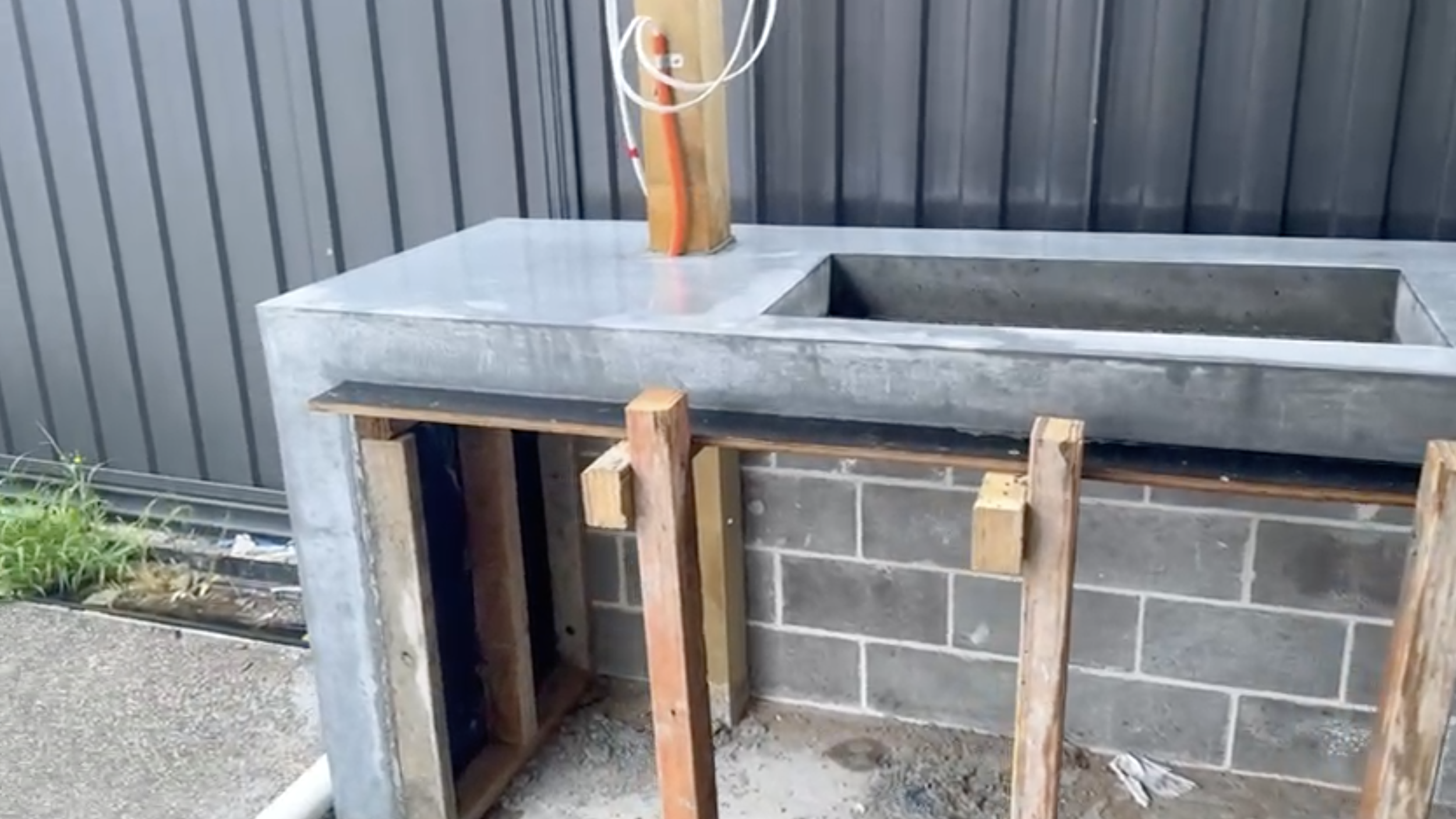 Outdoor concrete sink with wooden supports, next to a brick wall and a black metal fence