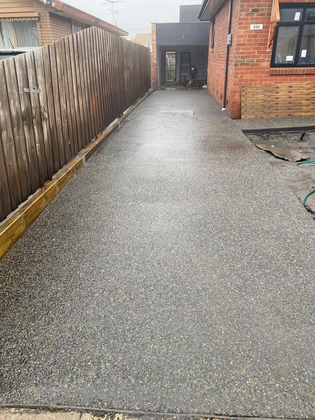 Freshly laid concrete driveway between a wooden fence and a brick house, with construction tools and materials in the background.