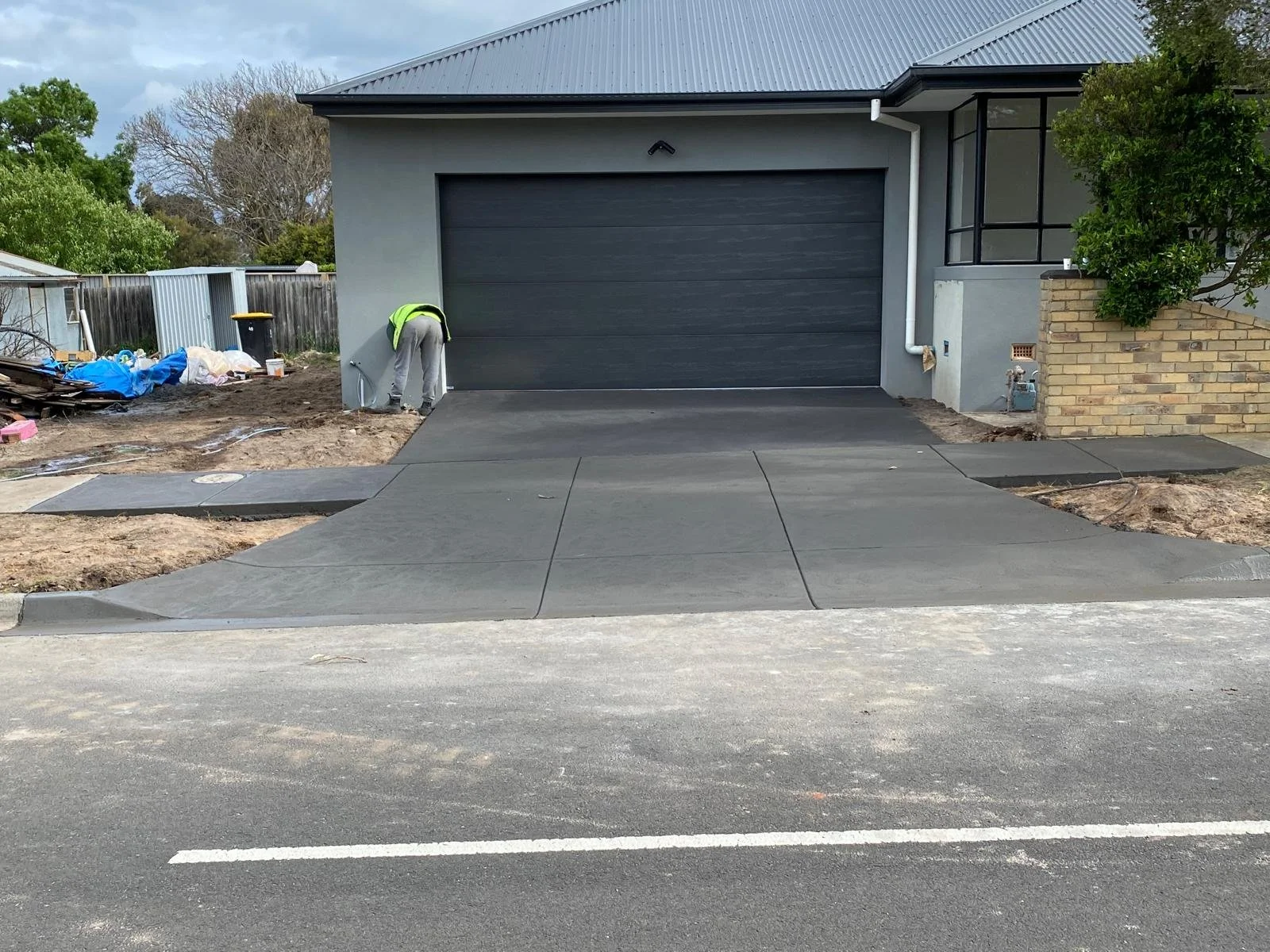 A person working on a newly paved driveway in front of a modern house with a dark garage door, brick and gray exterior walls, and a corner porch with glass panels.