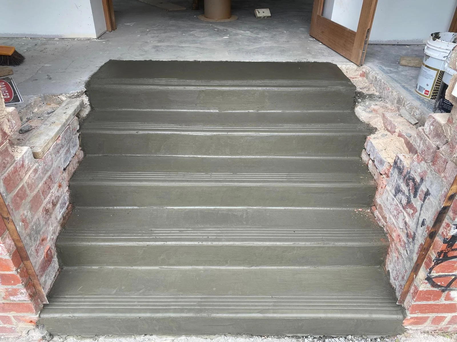 Newly poured concrete steps with textured lines for grip, adjacent to brick walls, leading to a doorway in a construction area.