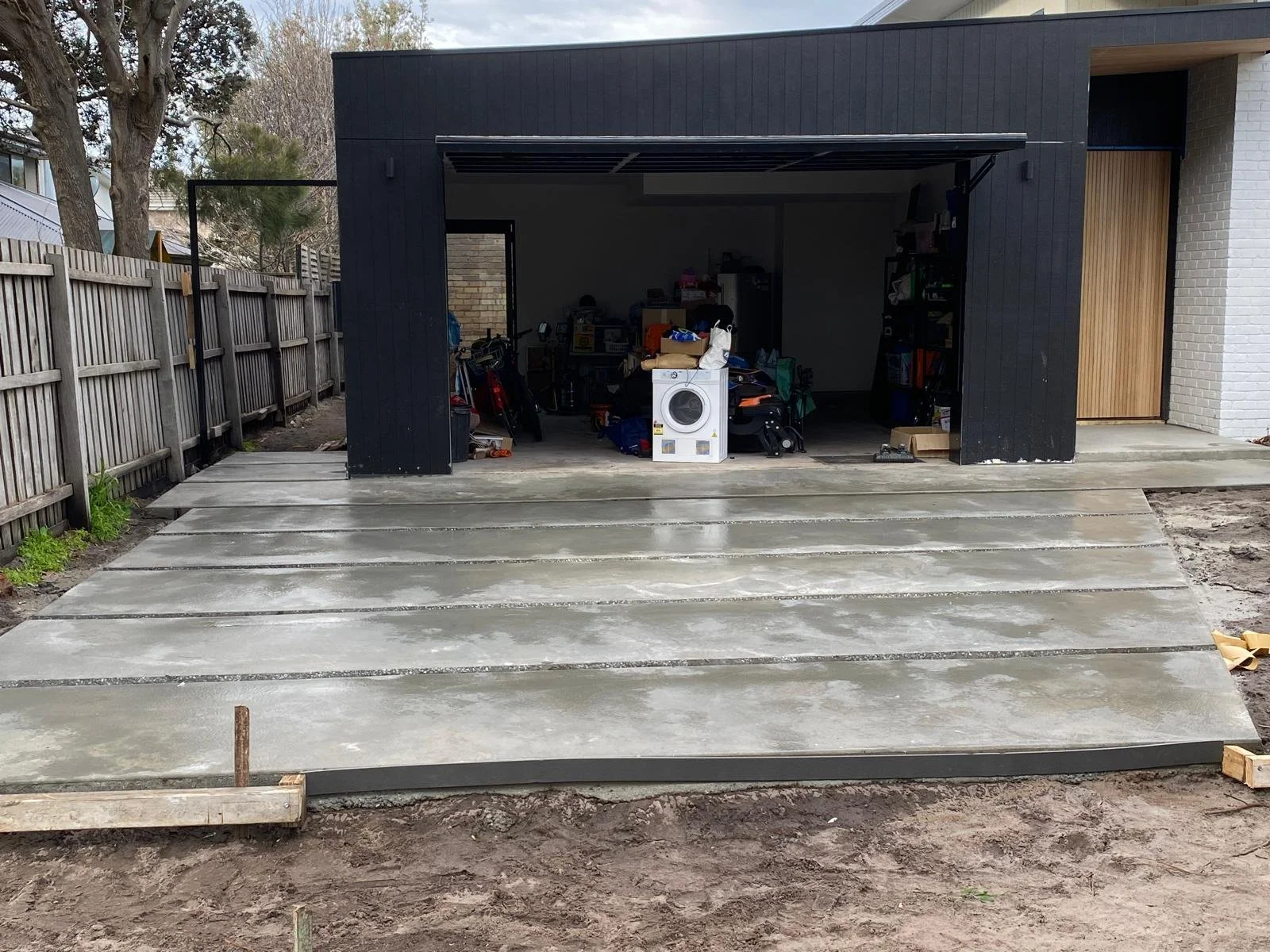 Concrete driveway leading to a garage with an open door, with various items stored inside, and a wooden fence on the left side.