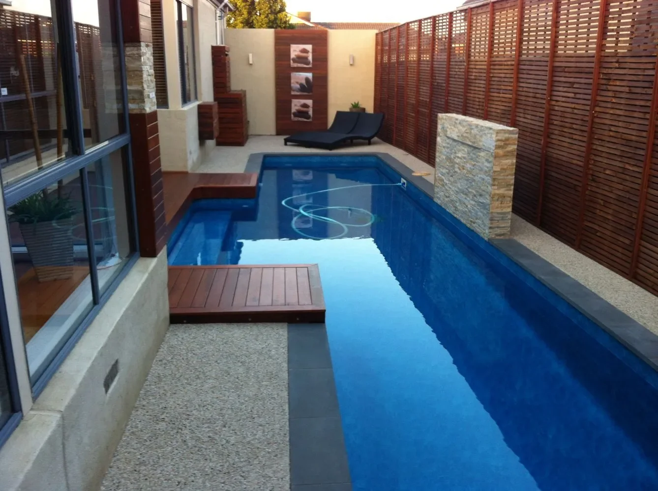 Modern backyard with a small rectangular swimming pool, wooden deck, black lounge chair, and decorative wall art, enclosed by a wooden fence.