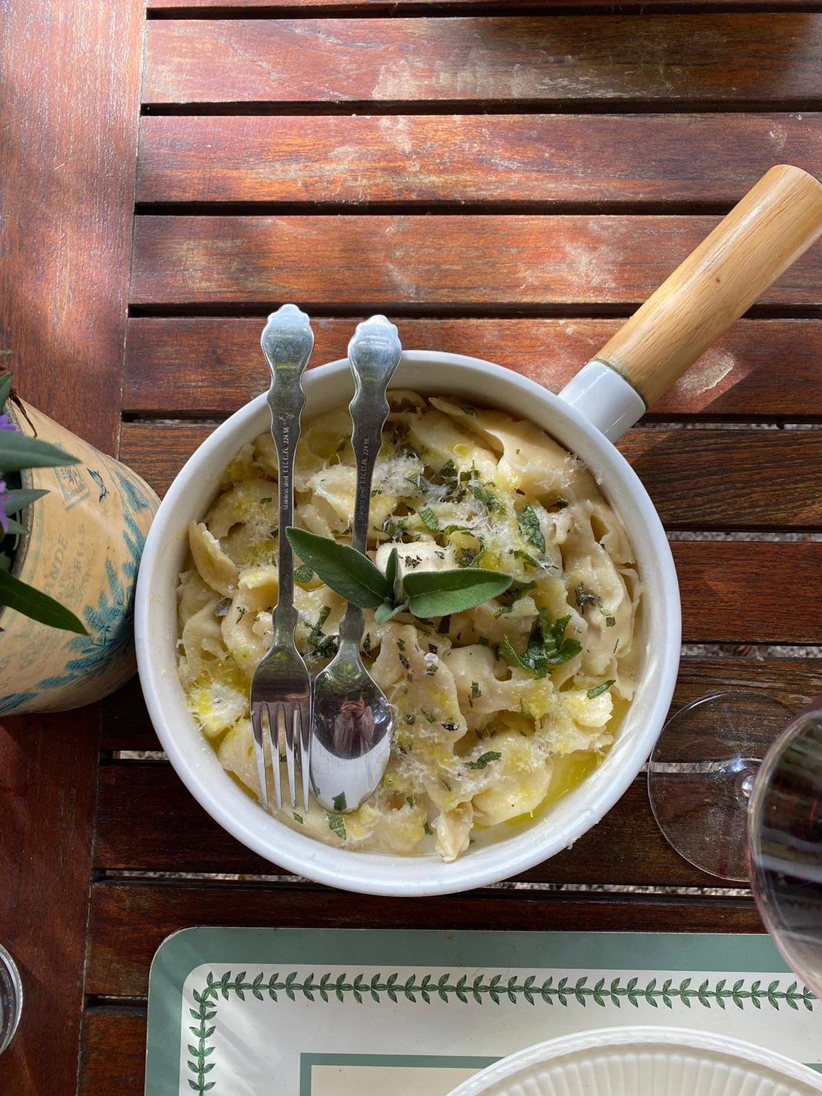 A bowl of pasta with cheese, herbs, and possibly a creamy sauce, garnished with a few olive leaves, on a wooden table with two metal forks resting in the bowl.