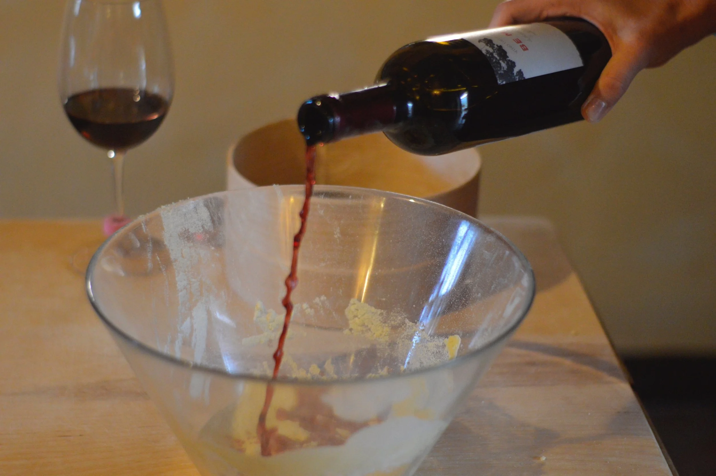 Pouring red wine into a glass bowl on a wooden table, with a full glass of red wine in the background.