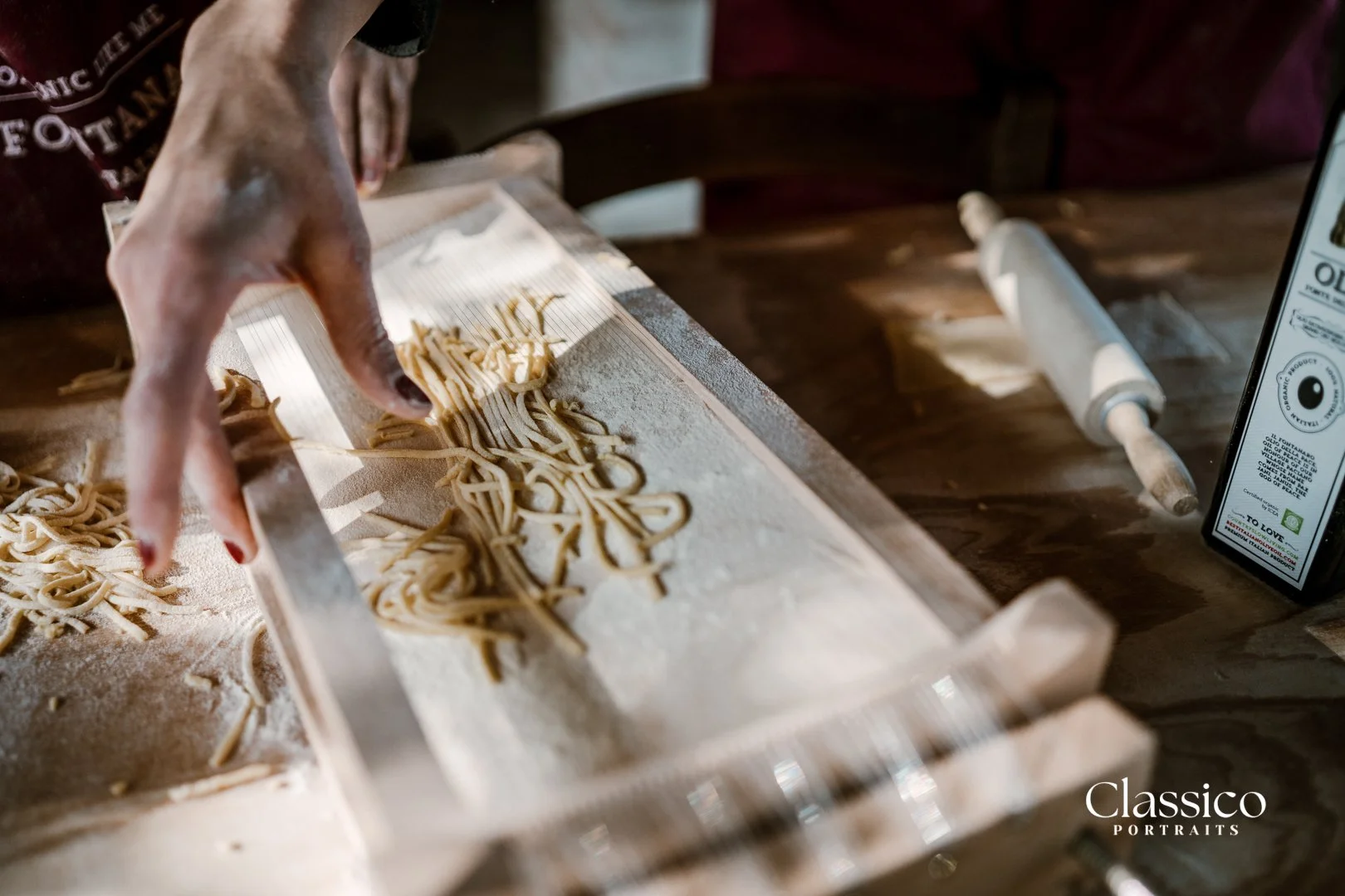 Hand cutting freshly made pasta on a floured wooden surface with a pasta cutter, a rolling pin, and a bottle of olive oil on a wooden table.
