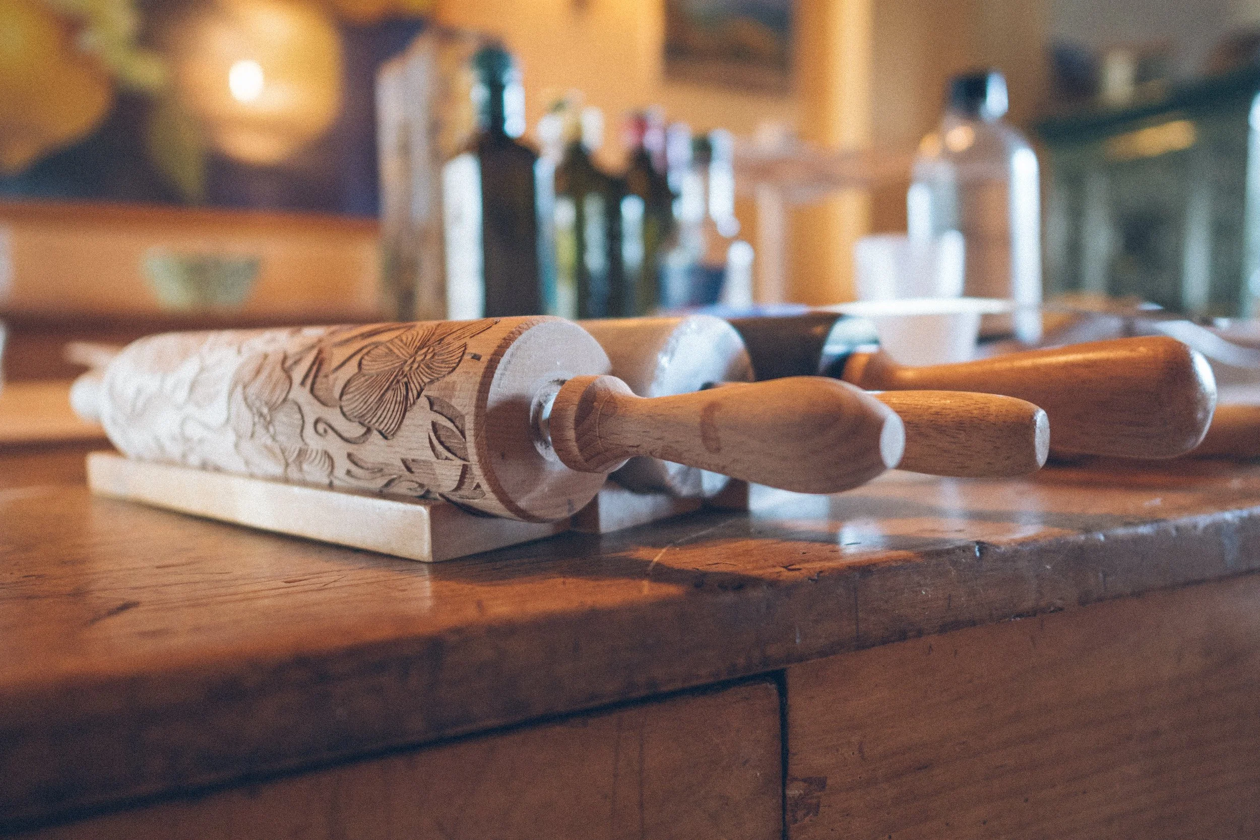 Wooden rolling pin with engraved floral design on a kitchen counter, with various bottles and kitchen tools blurred in the background.