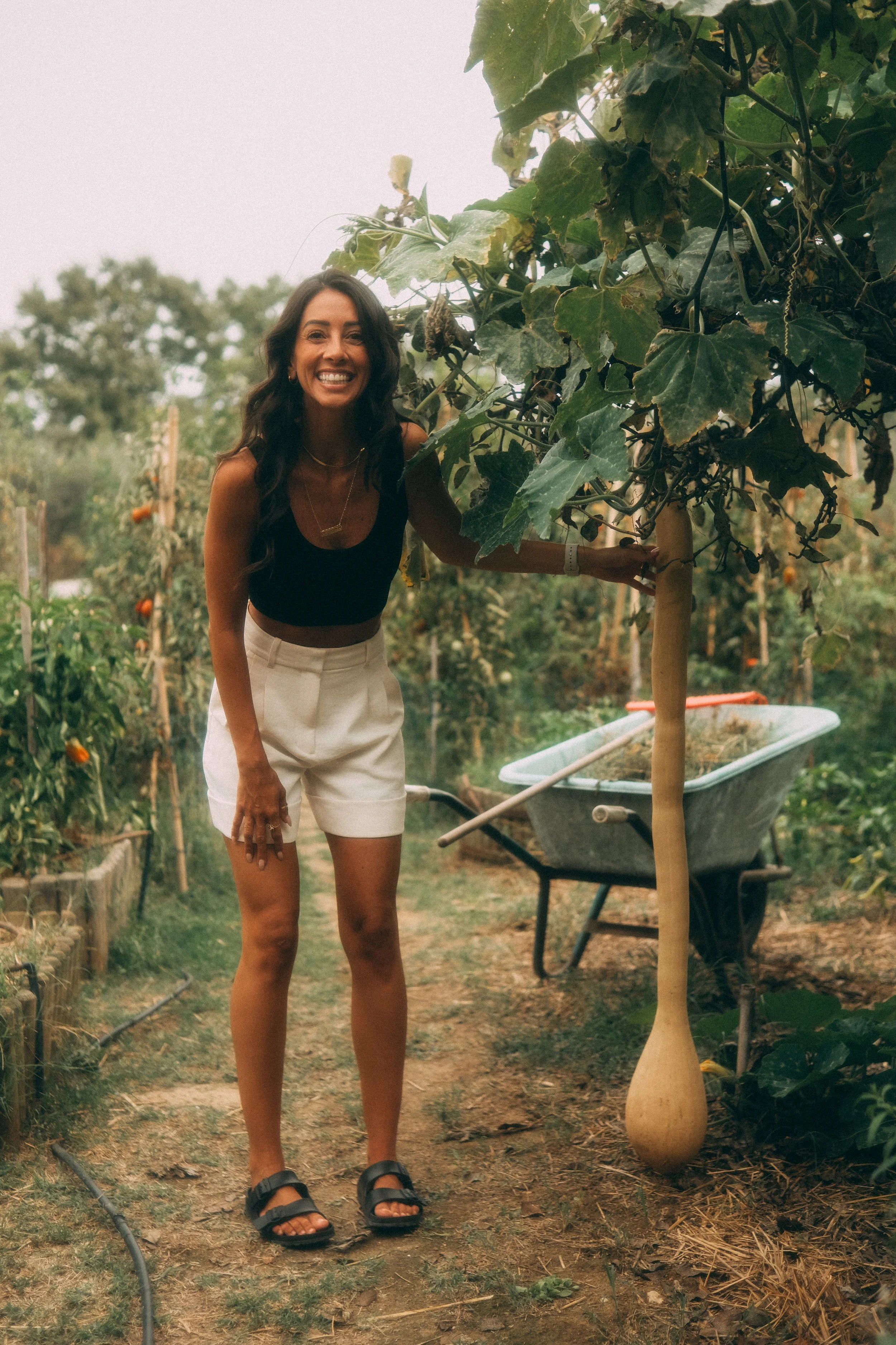 A woman smiling and standing in a garden or farm, holding a large gourd hanging from a vine. She wears a black crop top, beige shorts, and sandals, with a wheelbarrow nearby.